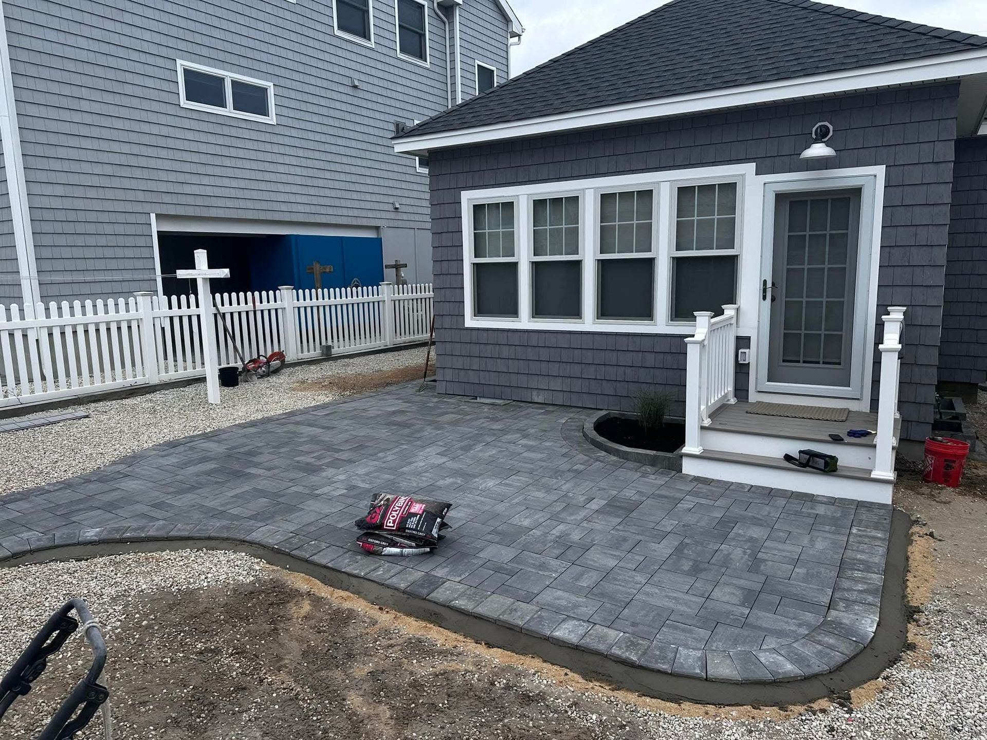 Grey paver patio in front of a grey house with white trim. A black border edges the patio.