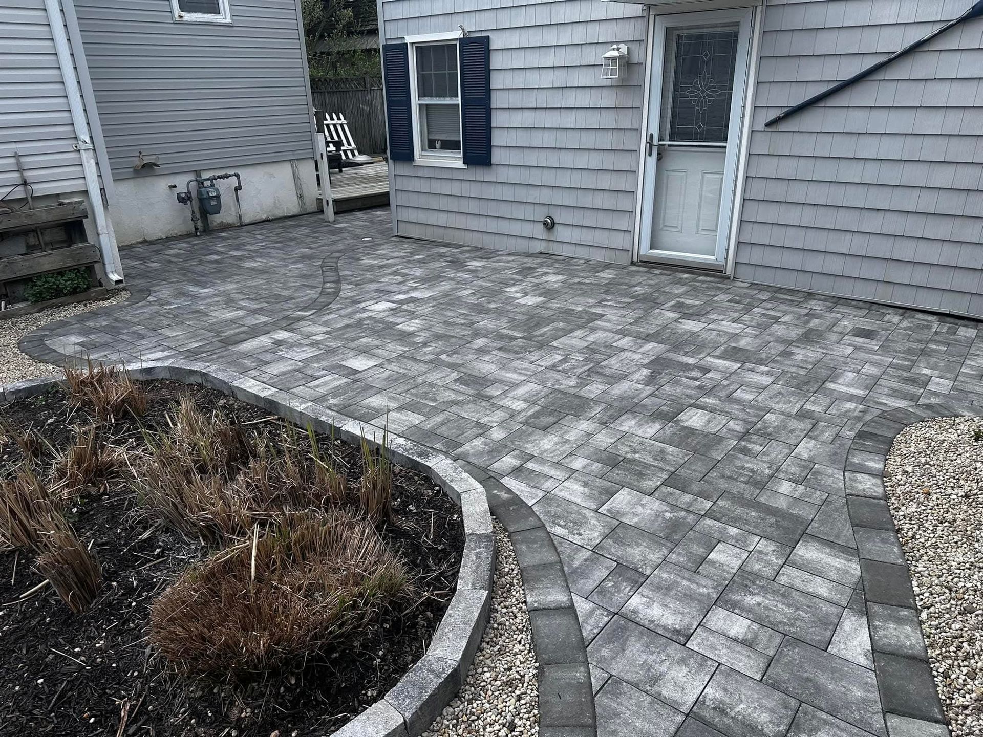 Paver patio with a bordering garden bed and pathway next to a gray building with a door.