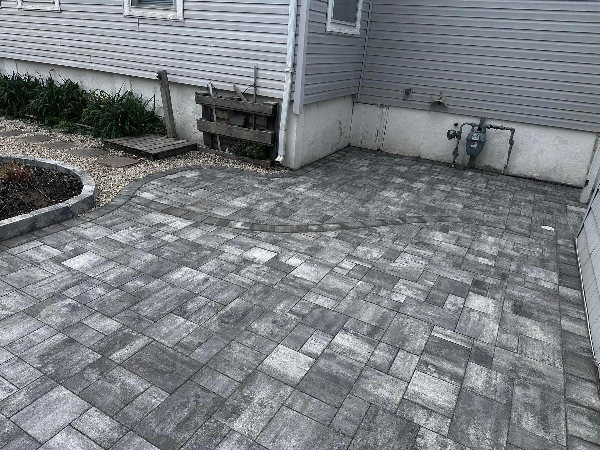 Grey paver patio next to a light gray house with gas meter.