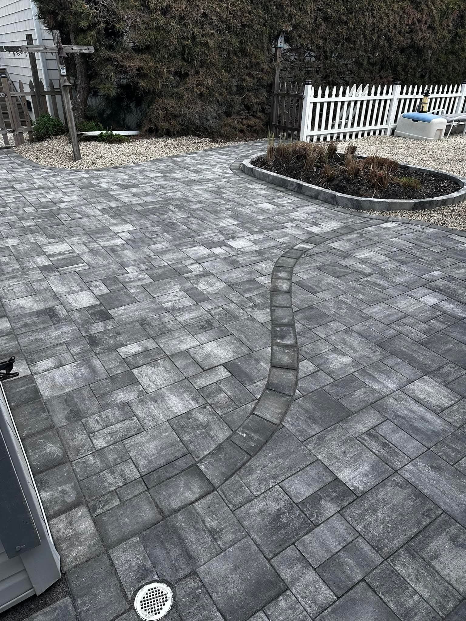 Brick patio with winding path, garden bed, white picket fence, and a dark green hedge.
