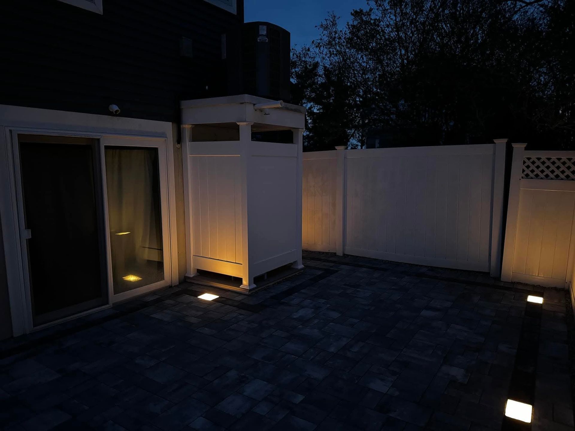 Nighttime outdoor patio with illuminated in-ground lights, white fence, and sliding glass door.
