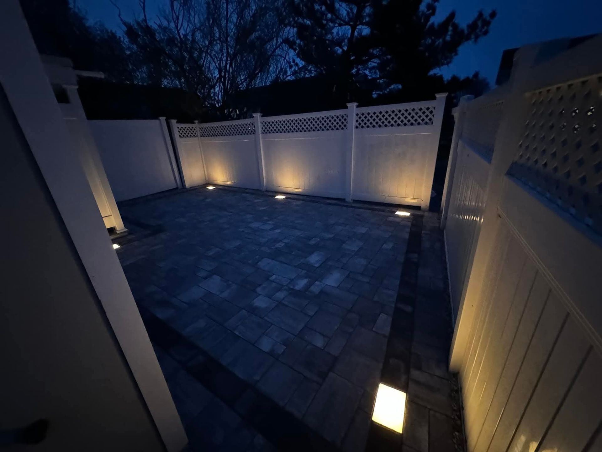 A dark backyard patio with illuminated pavers, white fence, and ambient lighting.