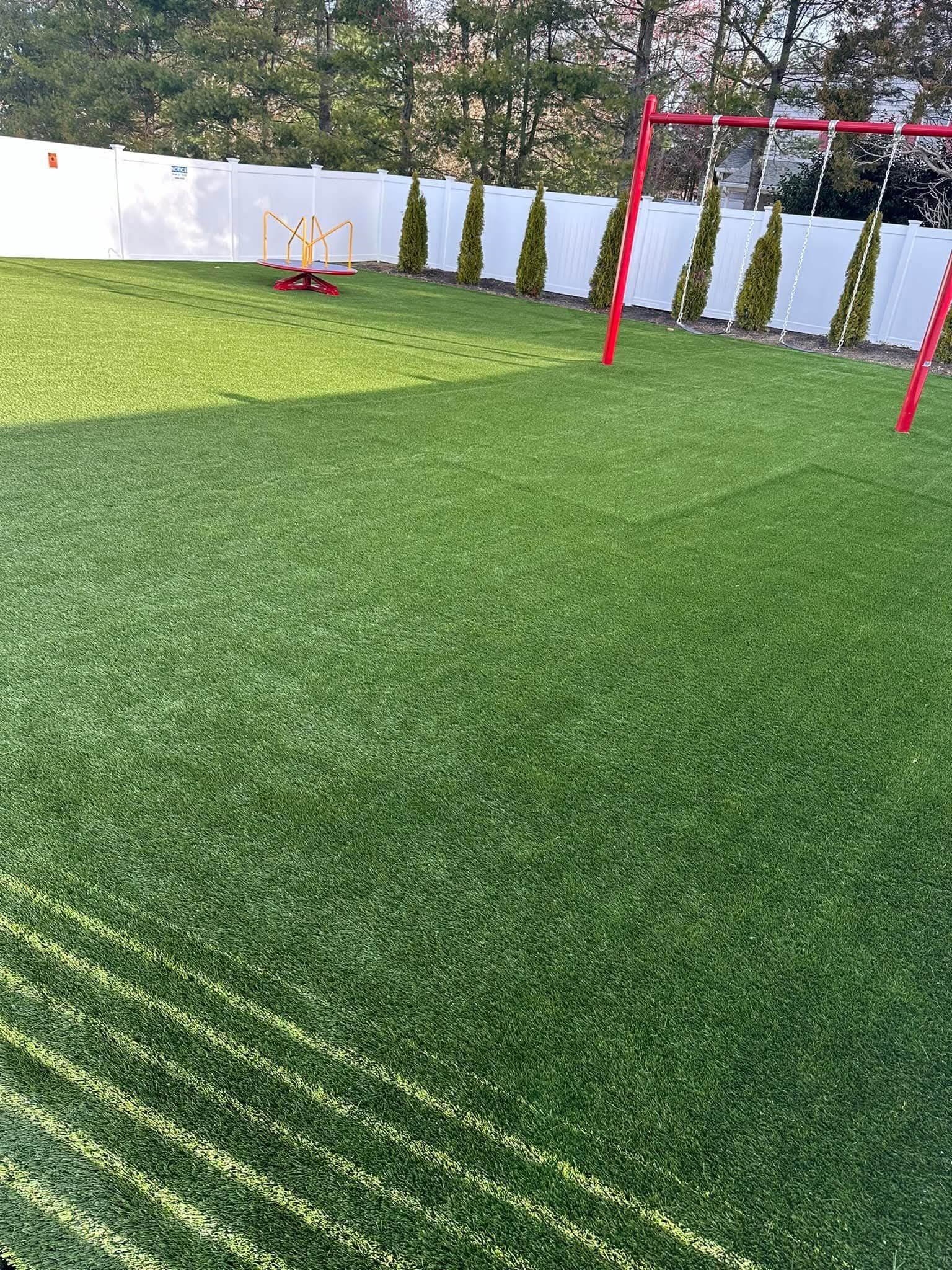 Green artificial turf playground with a red swing set and white fence.