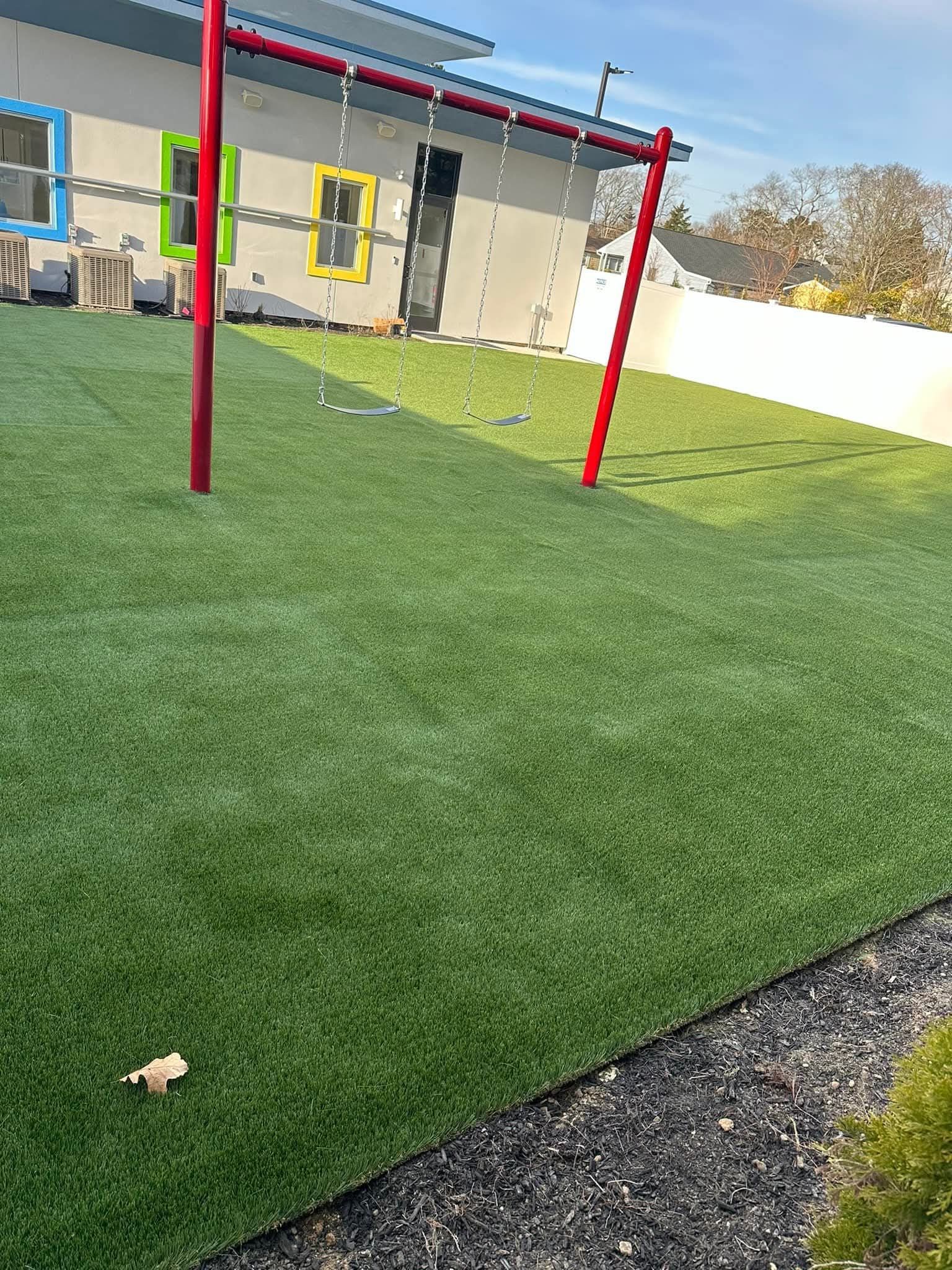 A green, artificial grass playground with a red swing set; building with colorful windows in the background.