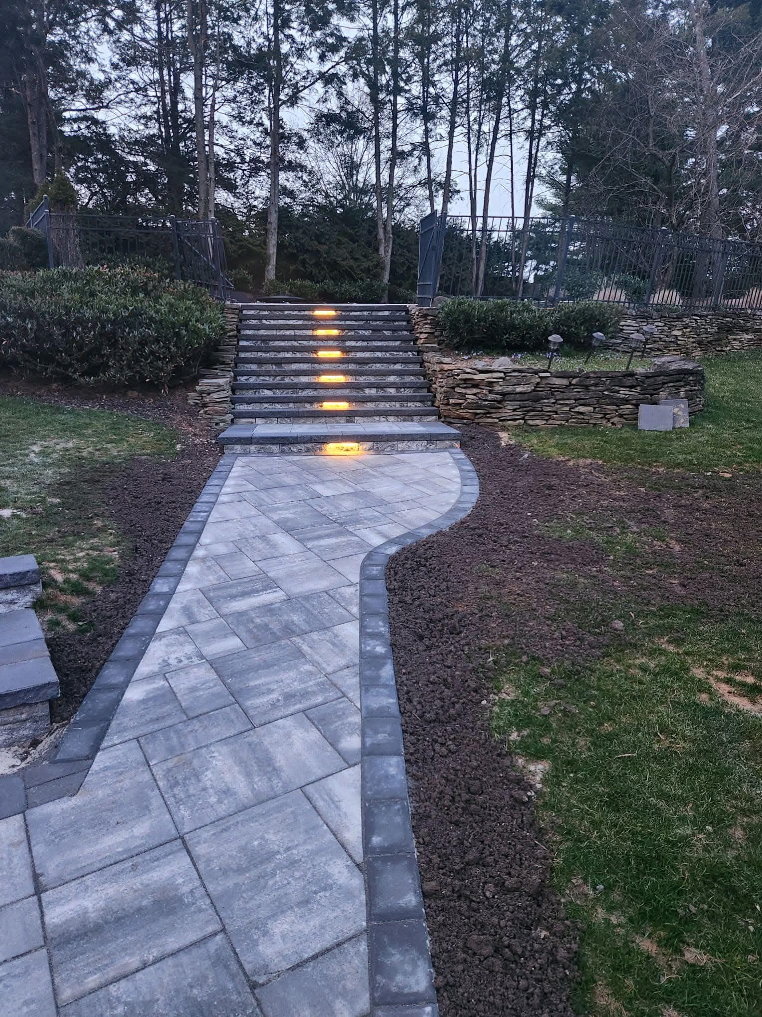 Paver pathway leading to lit steps and garden, dark gray bricks, outdoor setting.