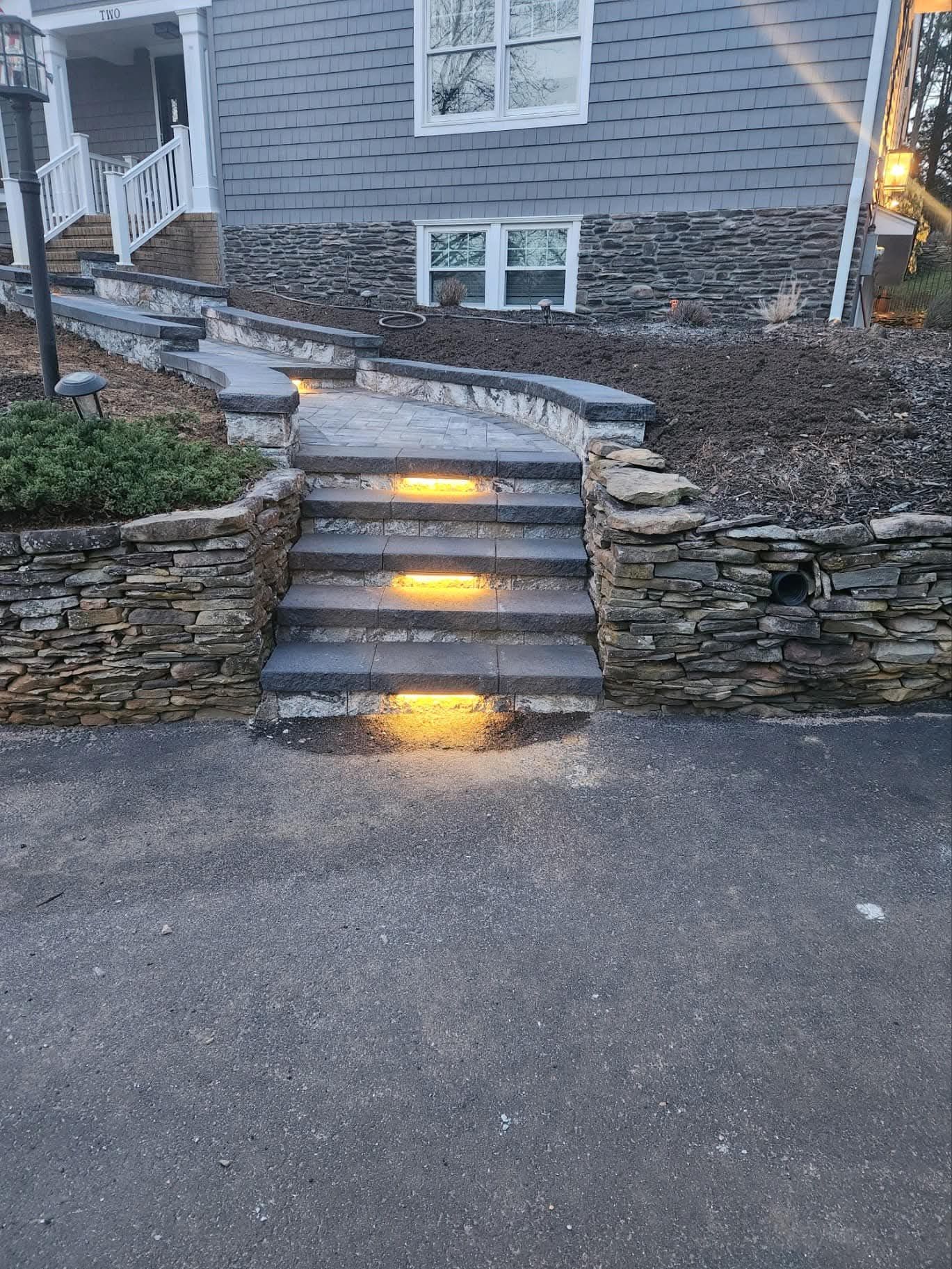 Stone steps with built-in lights leading to a house with stone and gray siding.