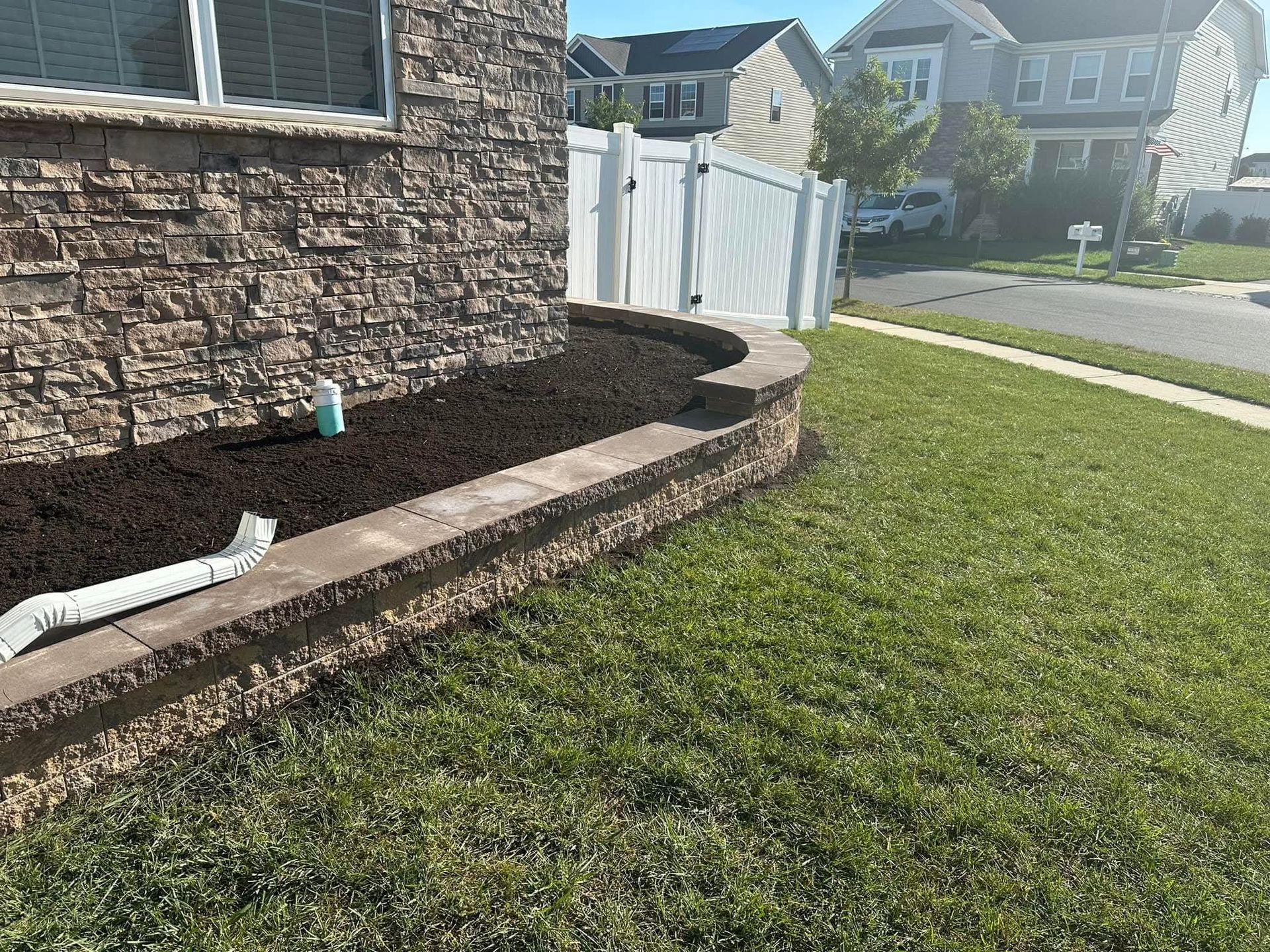 Brick wall garden bed with mulch, beside grass, in front of a house and a white fence.