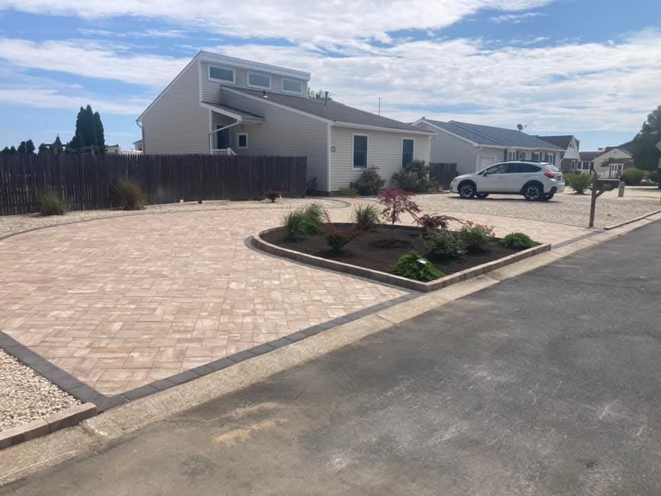 Paver driveway in front of a light-colored house, with a small garden and a car.