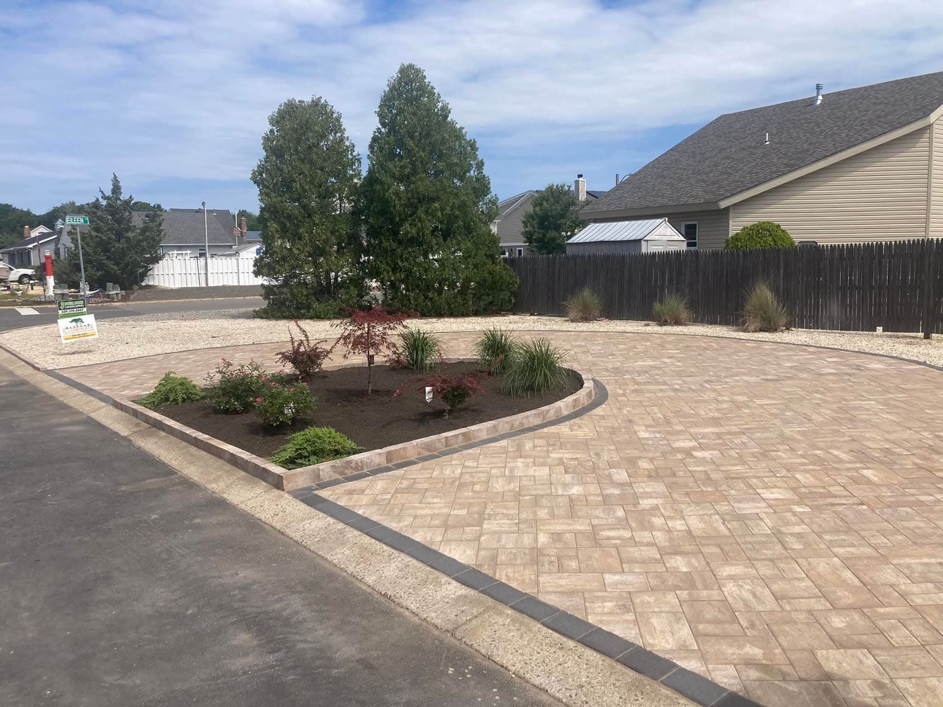 Brick paved driveway with garden bed and two tall trees against a residential setting.