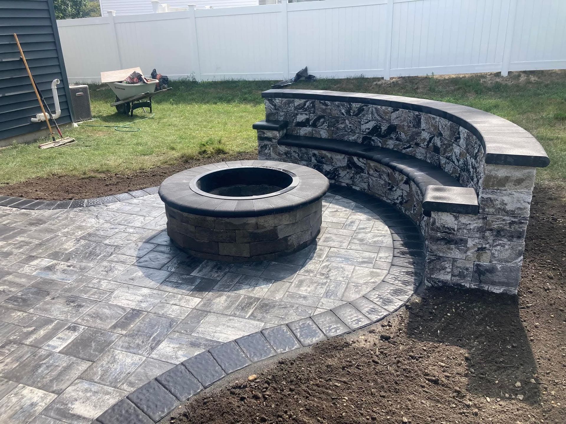 Fire pit with curved stone bench on a paved patio in a backyard setting.