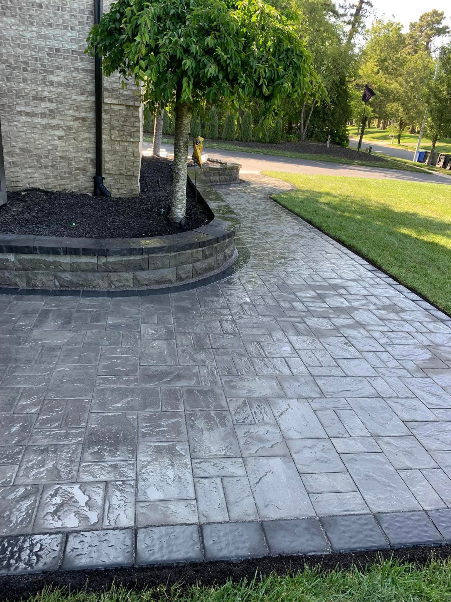 Stone walkway next to a curved planter and tree, adjacent to a grassy lawn and asphalt road.