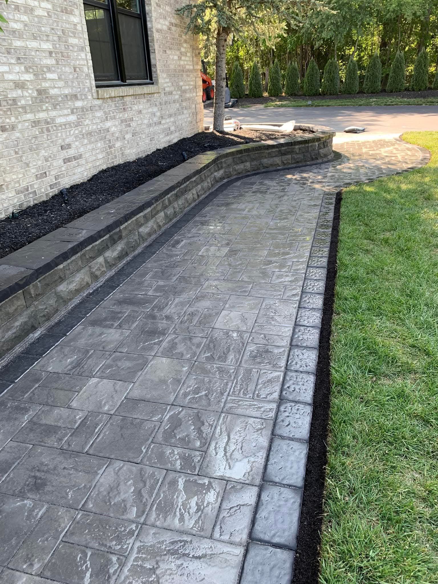 Brick walkway alongside grass and a building, with a retaining wall and black mulch.