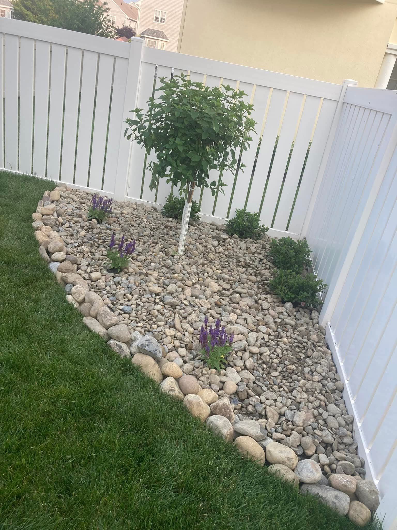 Landscaped backyard bed with rocks, border stones, a small tree, and purple flowers, by a white fence.