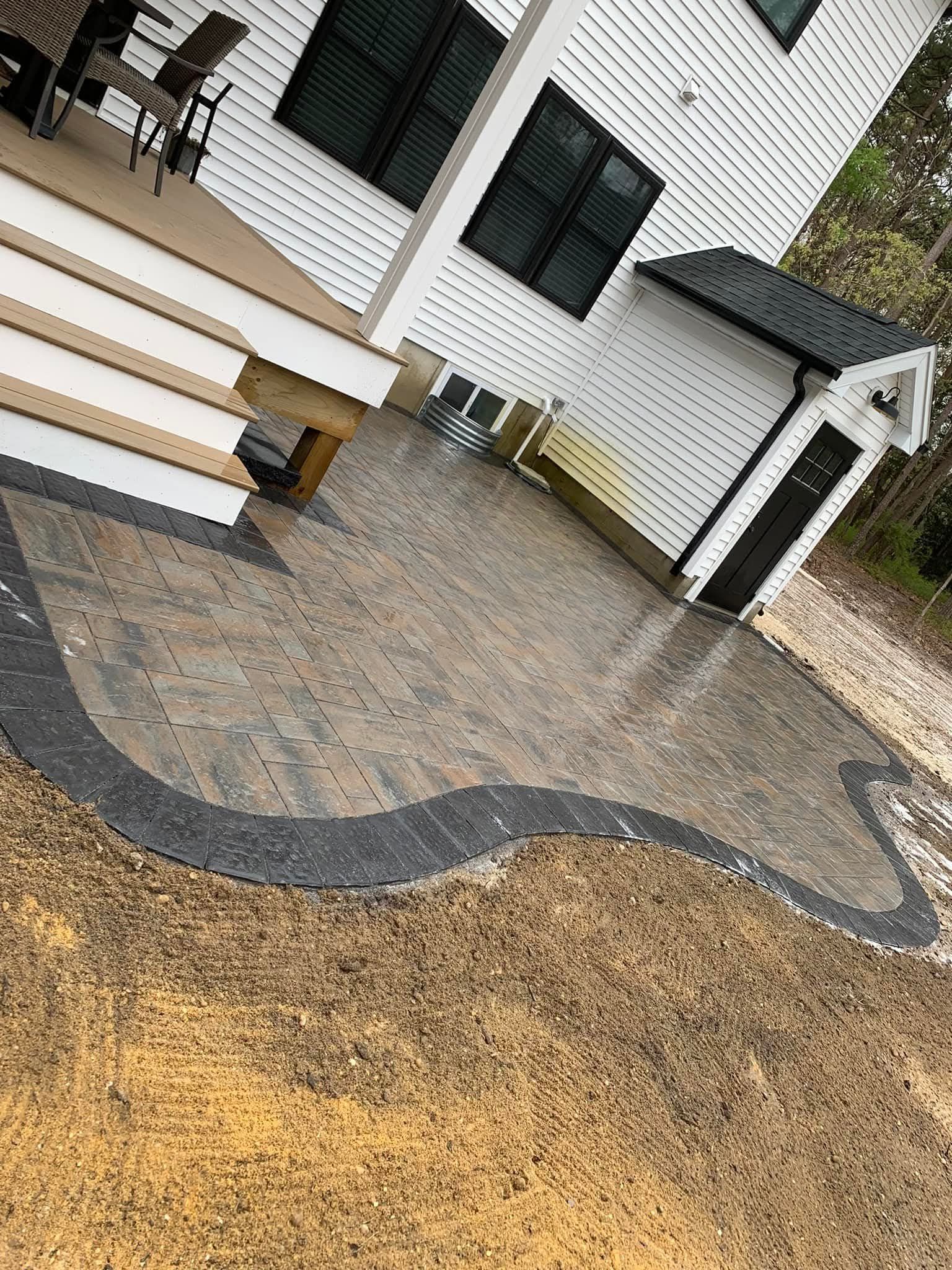 Backyard patio with patterned pavers, outlined by dark rocks, next to a white house.