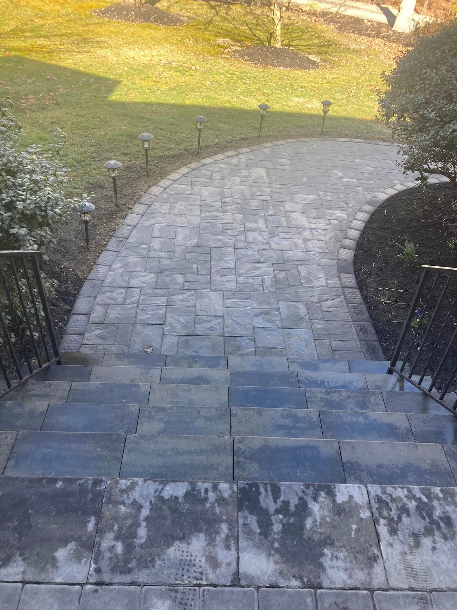 Stone walkway and steps leading to a garden path, framed by railings and shrubbery, in an outdoor setting.