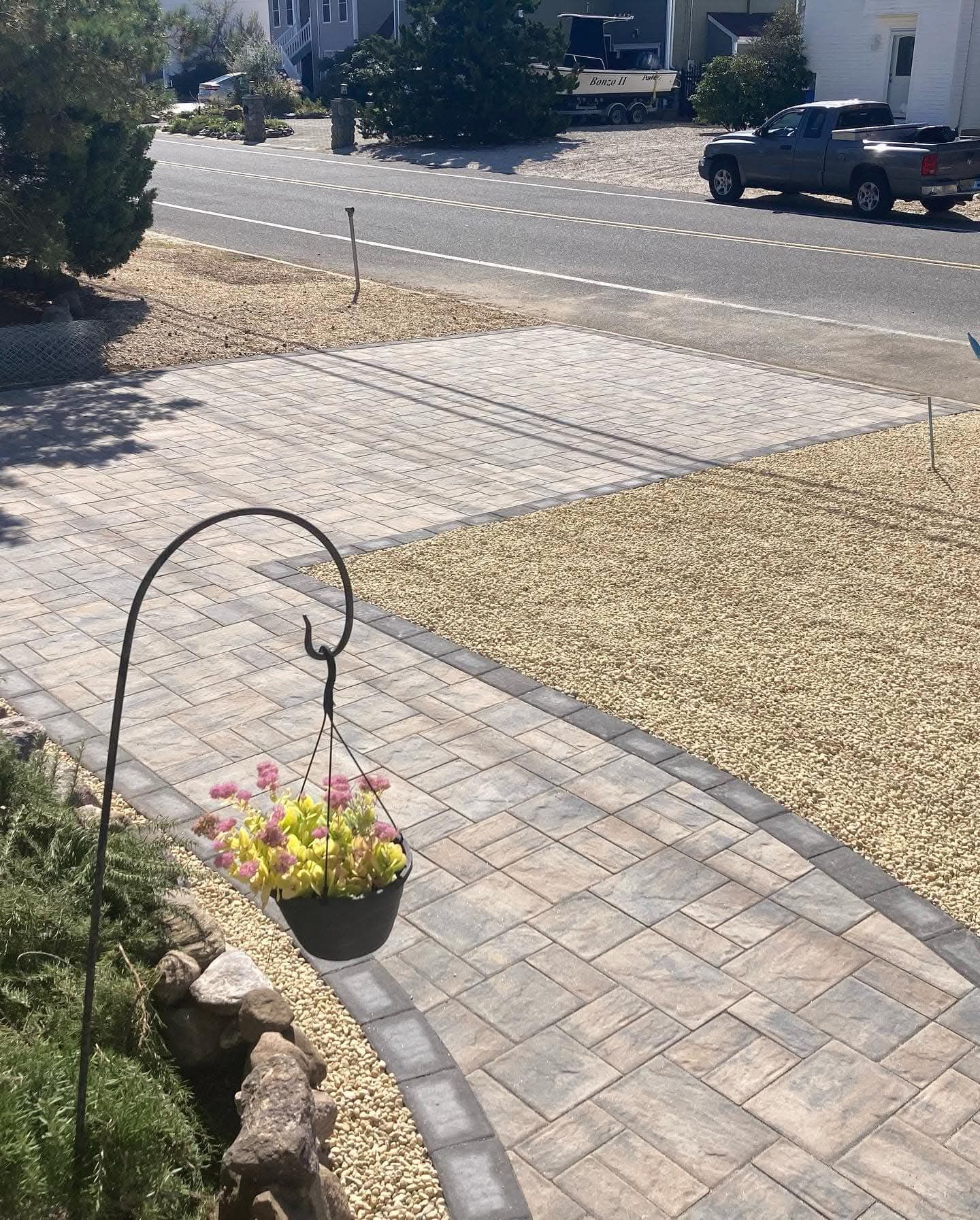 A brick walkway with a hanging flower basket leads to a street with a car driving by.