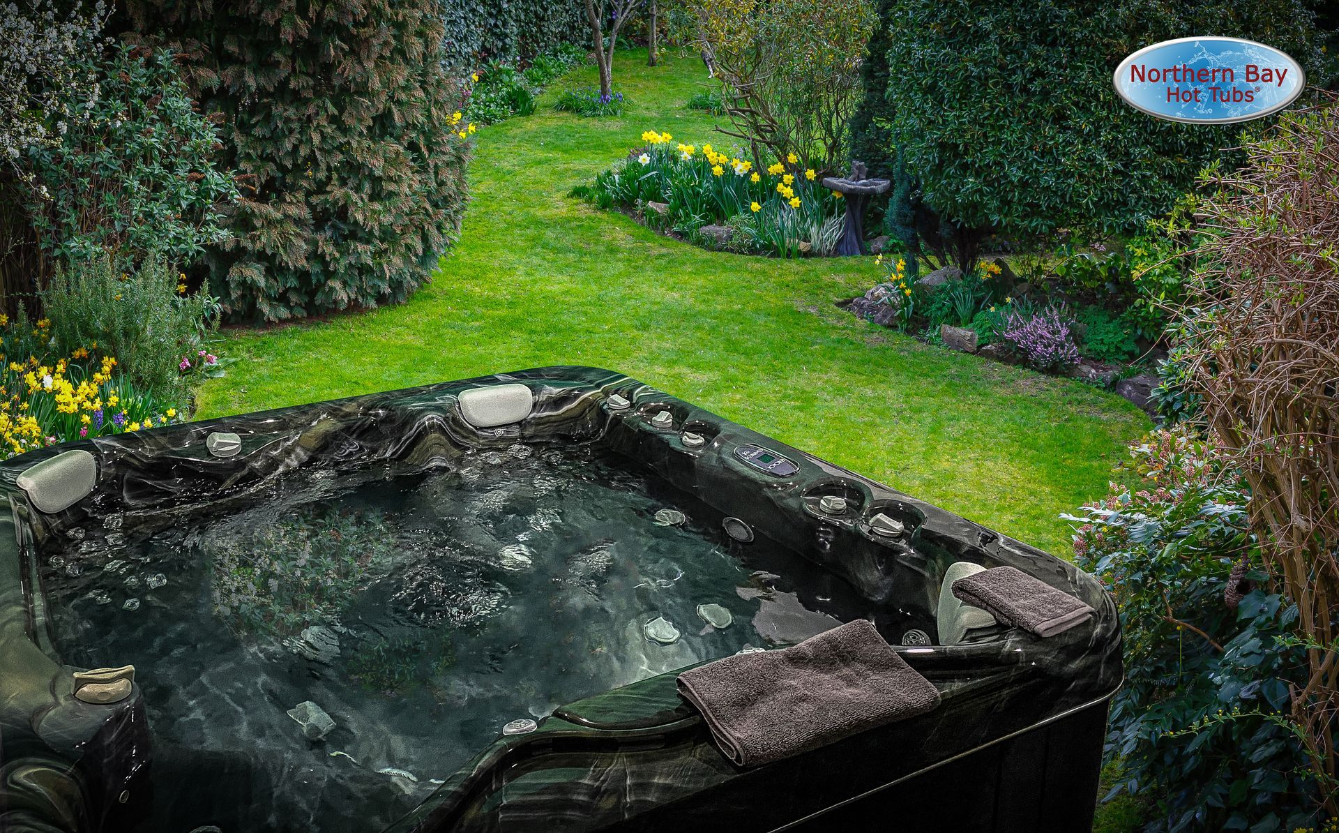 Hot tub in a lush green garden setting, surrounded by trees and flowers.