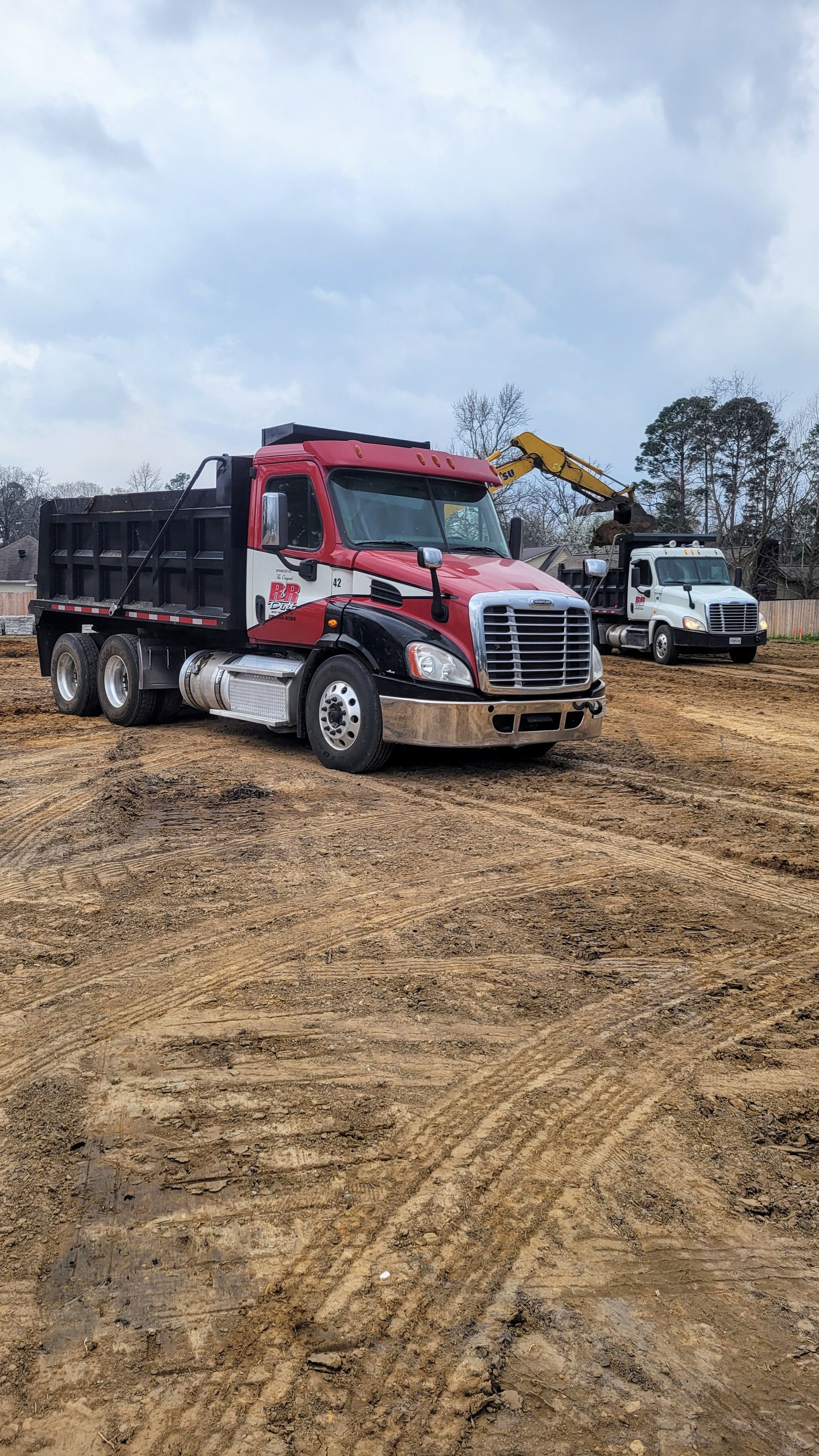 A red and black dump truck on a dirt field, with a construction vehicle loading it.