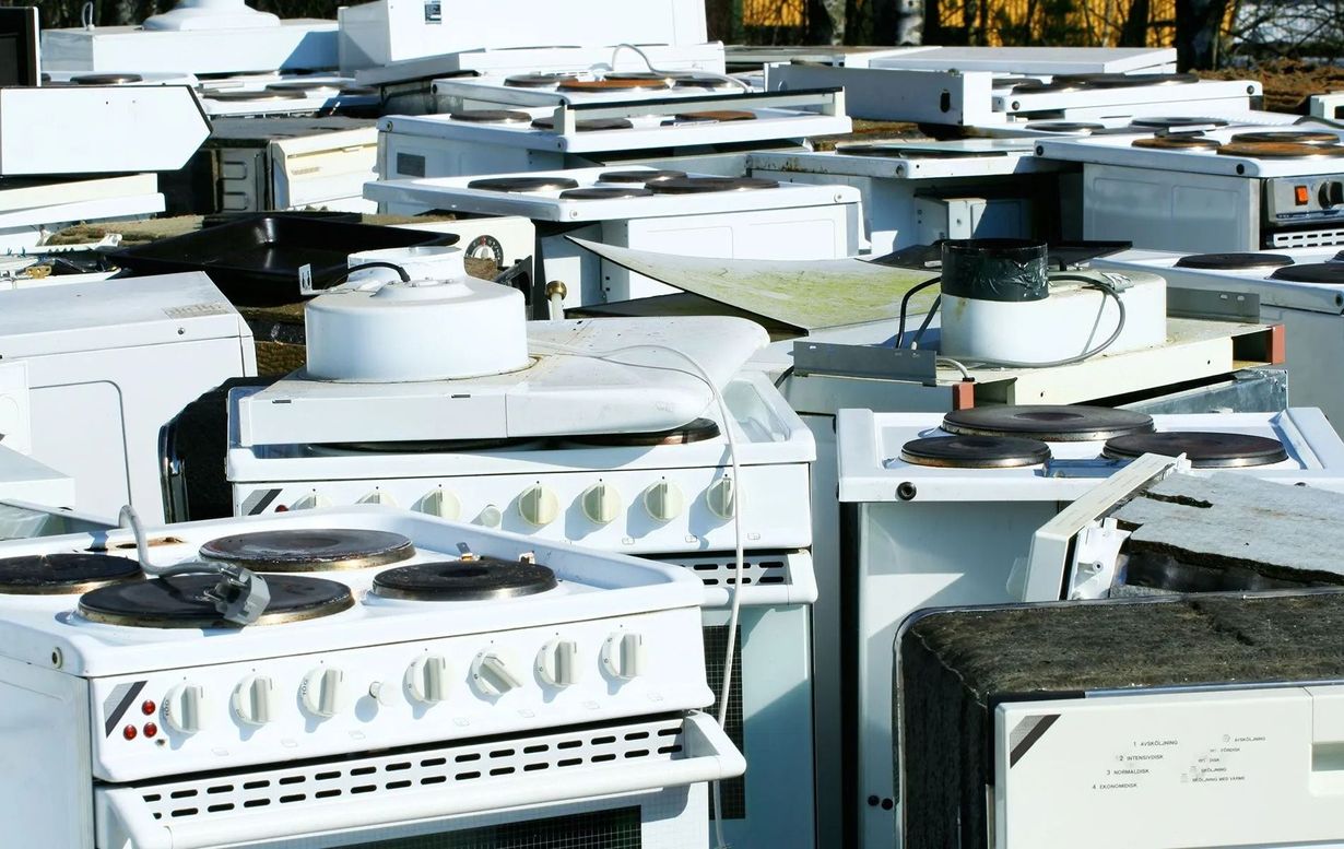 Pile of discarded white kitchen appliances in an outdoor junkyard setting.