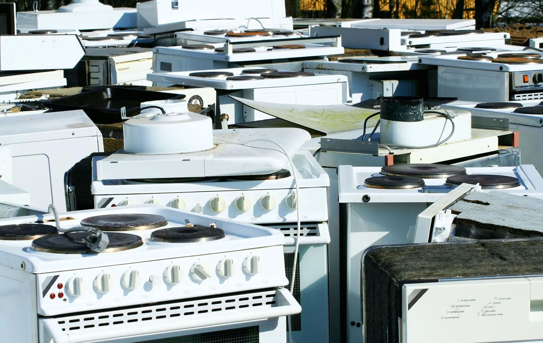 Pile of discarded white kitchen appliances in an outdoor junkyard setting.