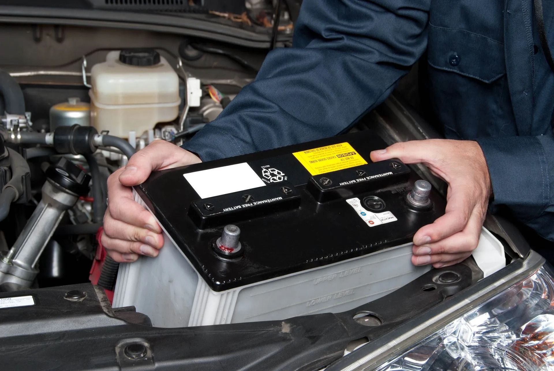 Mechanic holding a car battery in an engine bay, preparing to replace it.