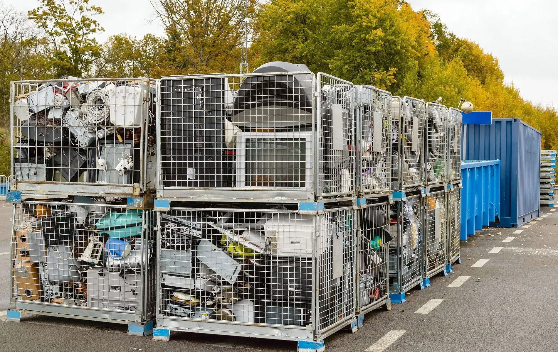 Cages filled with electronic waste, stacked outdoors, near a blue container and trees.