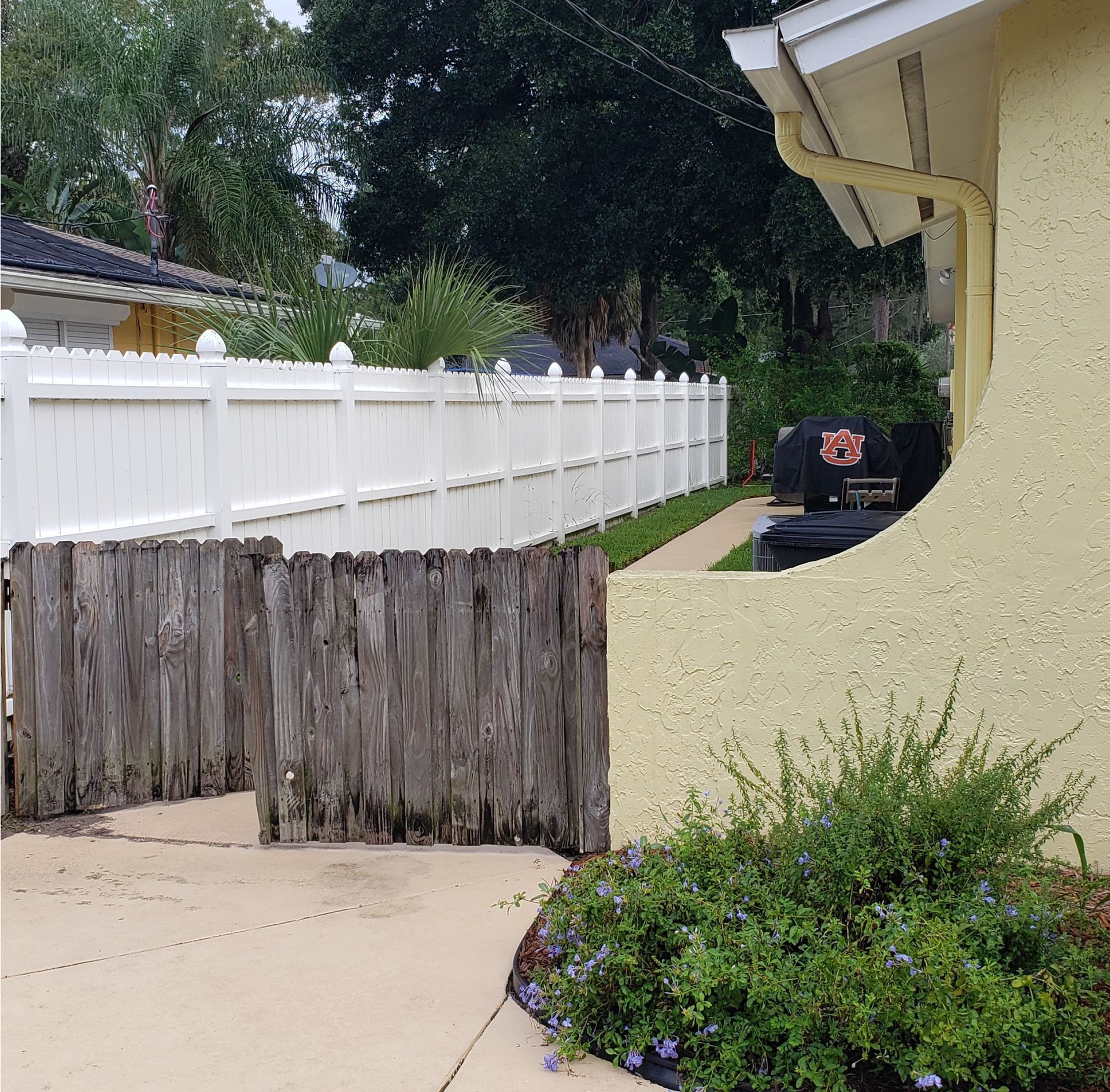 Driveway with weathered wooden gate, white fence, and a parked black vehicle beside a yellow house.