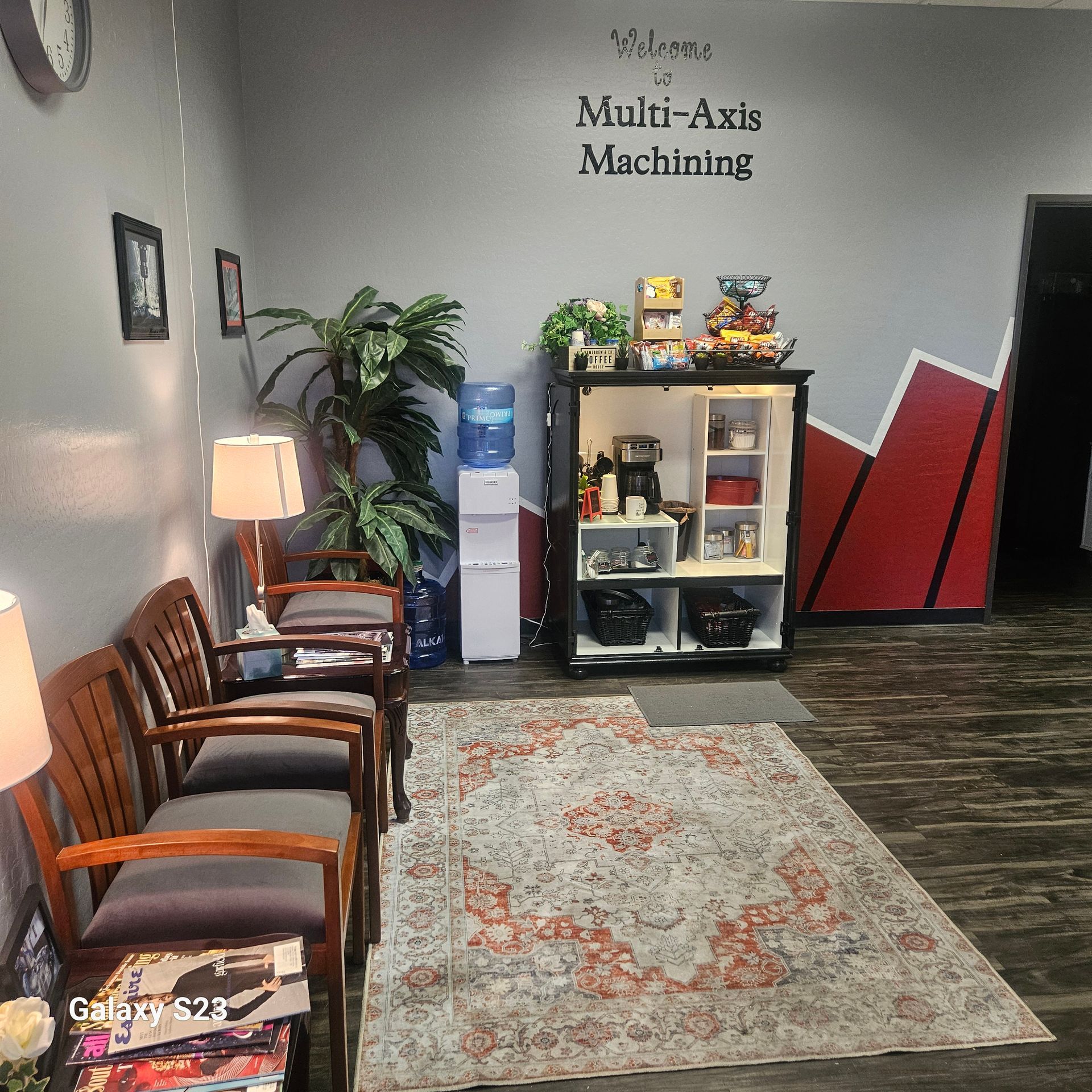 Waiting area for Multi-Axis Machining with wooden chairs, a water cooler, and a refreshment cabinet on a patterned rug.