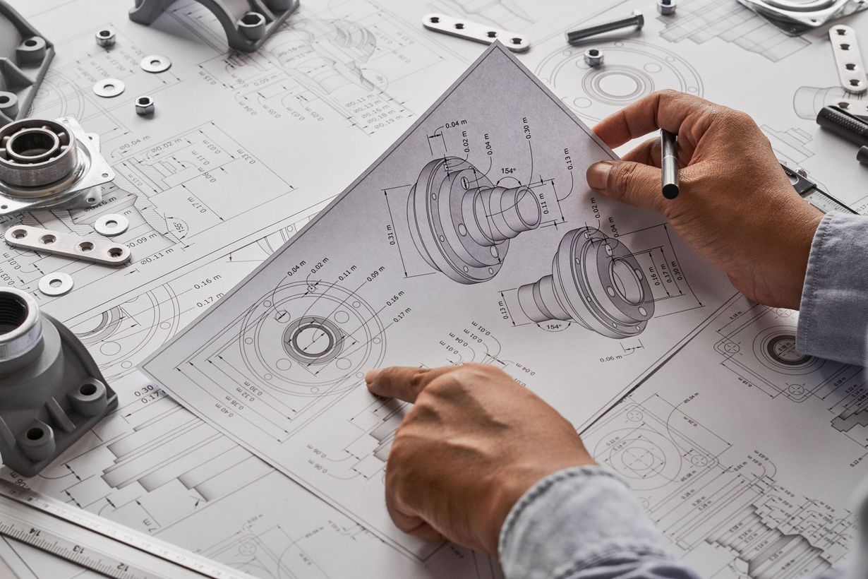An engineer examines technical blueprints for mechanical parts, with various metal components scattered on the workspace.