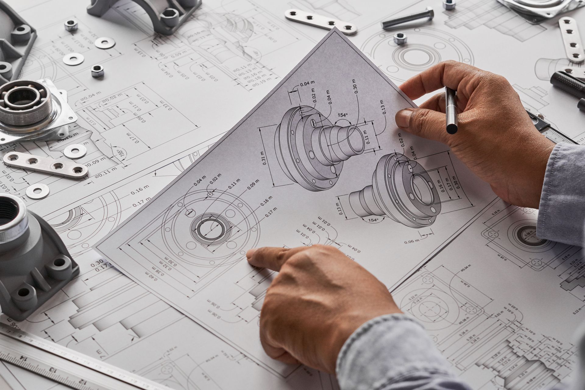 An engineer examines technical blueprints for mechanical parts, with various metal components scattered on the workspace.