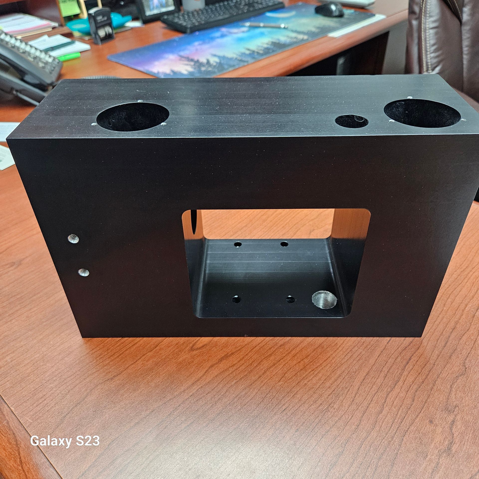 A black, rectangular mechanical component with circular and square cutouts, resting on a brown wooden desk.