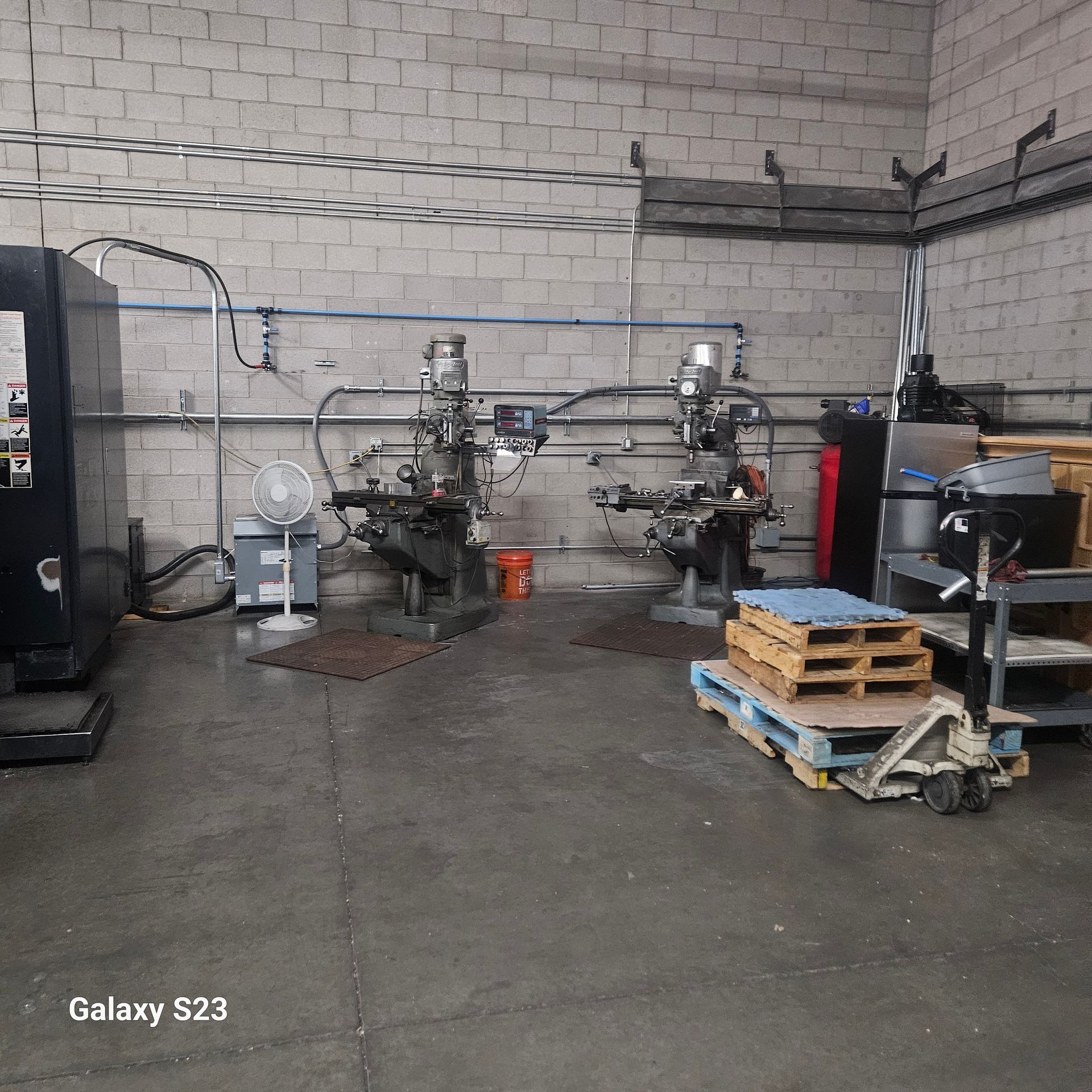 Industrial workshop featuring two milling machines, a floor fan, wooden pallets, and a hand truck against a brick wall.