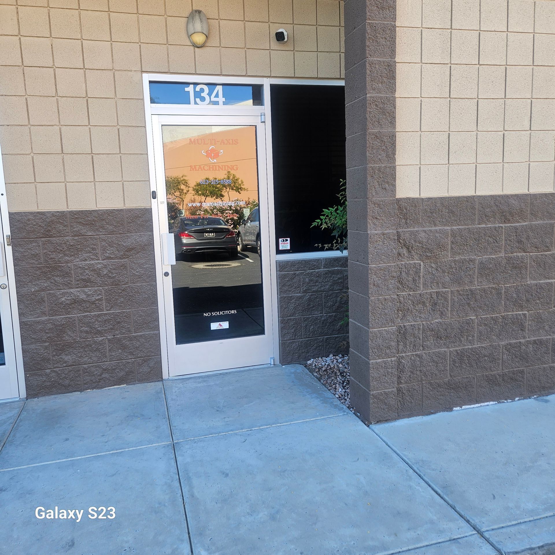 The entrance to unit 134 of a building with tan and dark brown masonry walls and a glass door reflecting a parking lot.