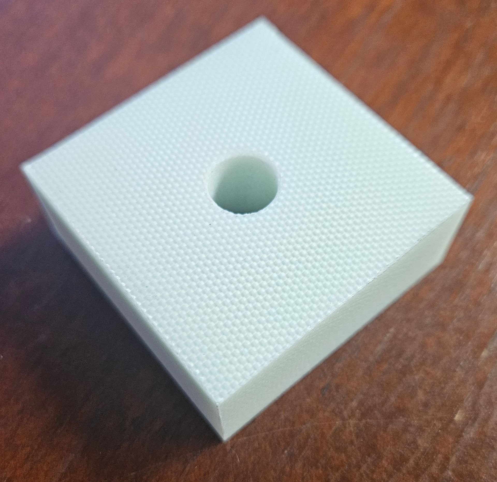 A light-colored, square plastic component with a fine, textured grid pattern on its surface and a circular hole in the center.