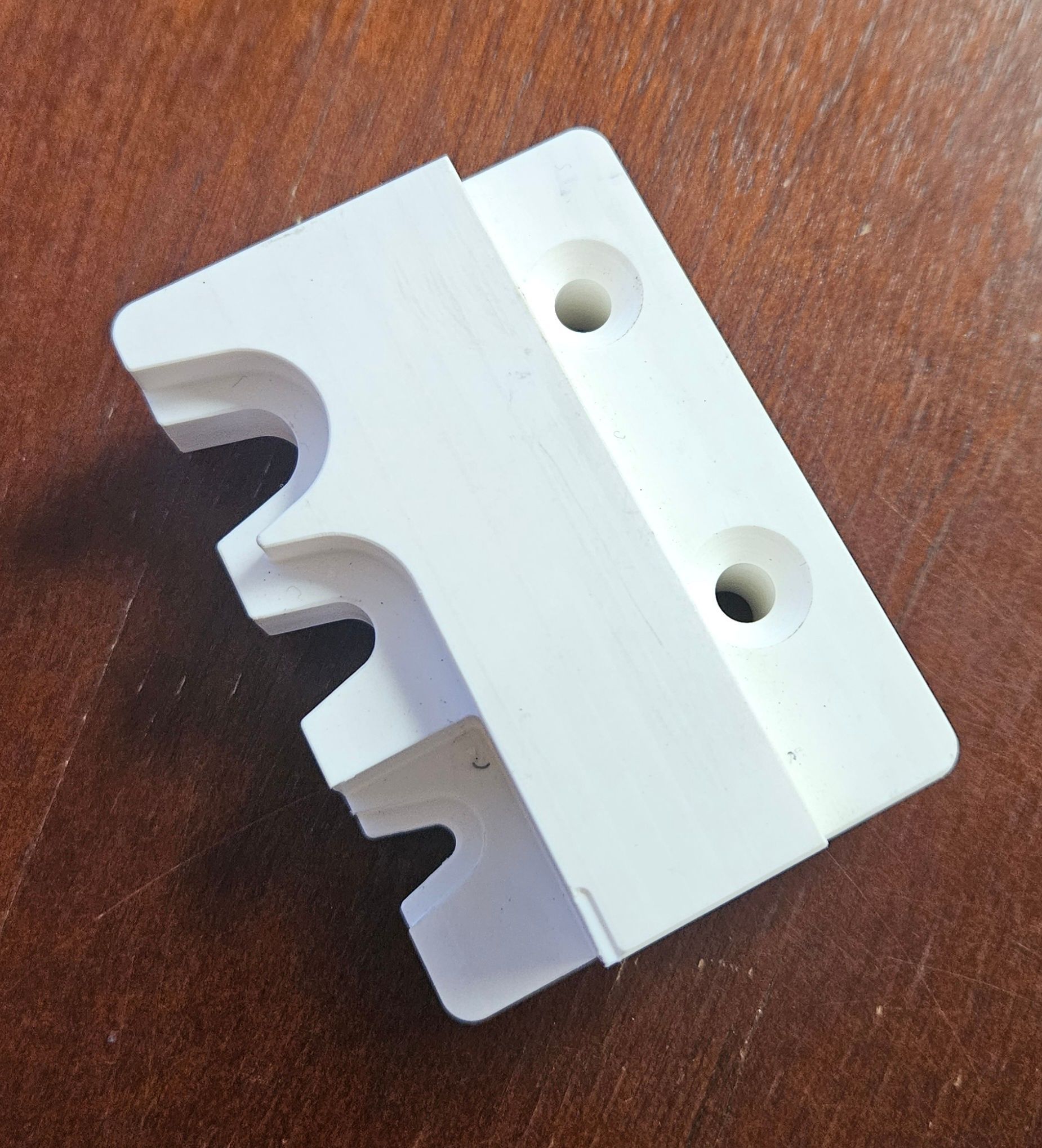 A white, rectangular plastic component with three semi-circular cutouts on one edge and two screw holes on the flat base.