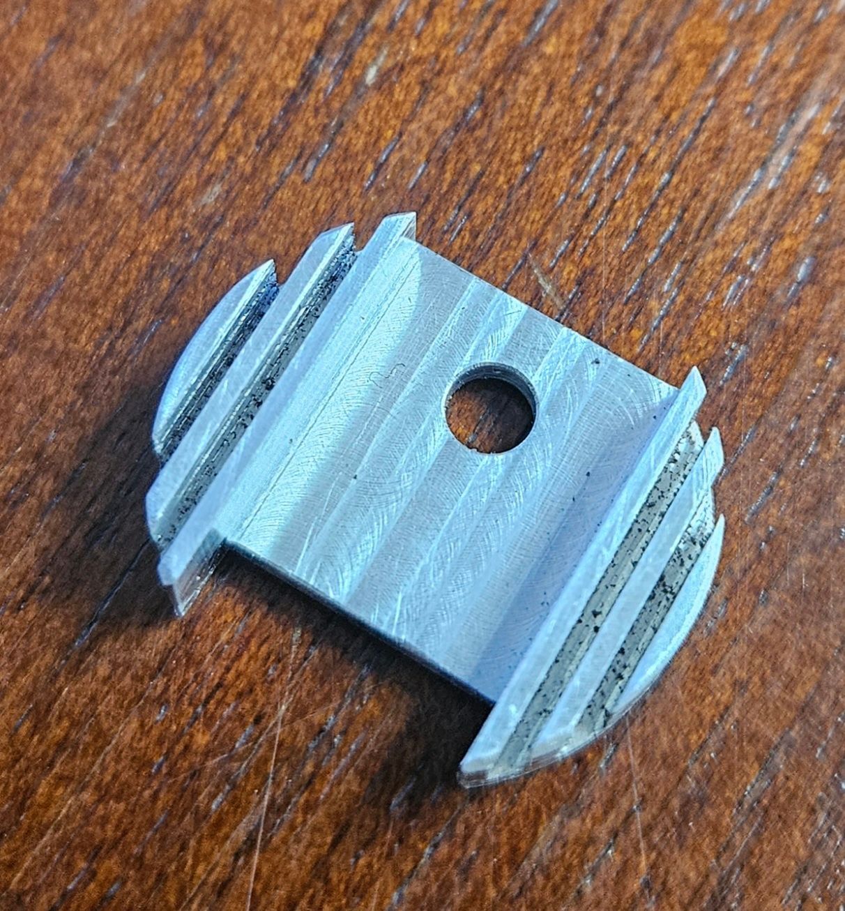 A small, rectangular, silver-colored metal piece with a circular hole in the center and finned edges, resting on wood.
