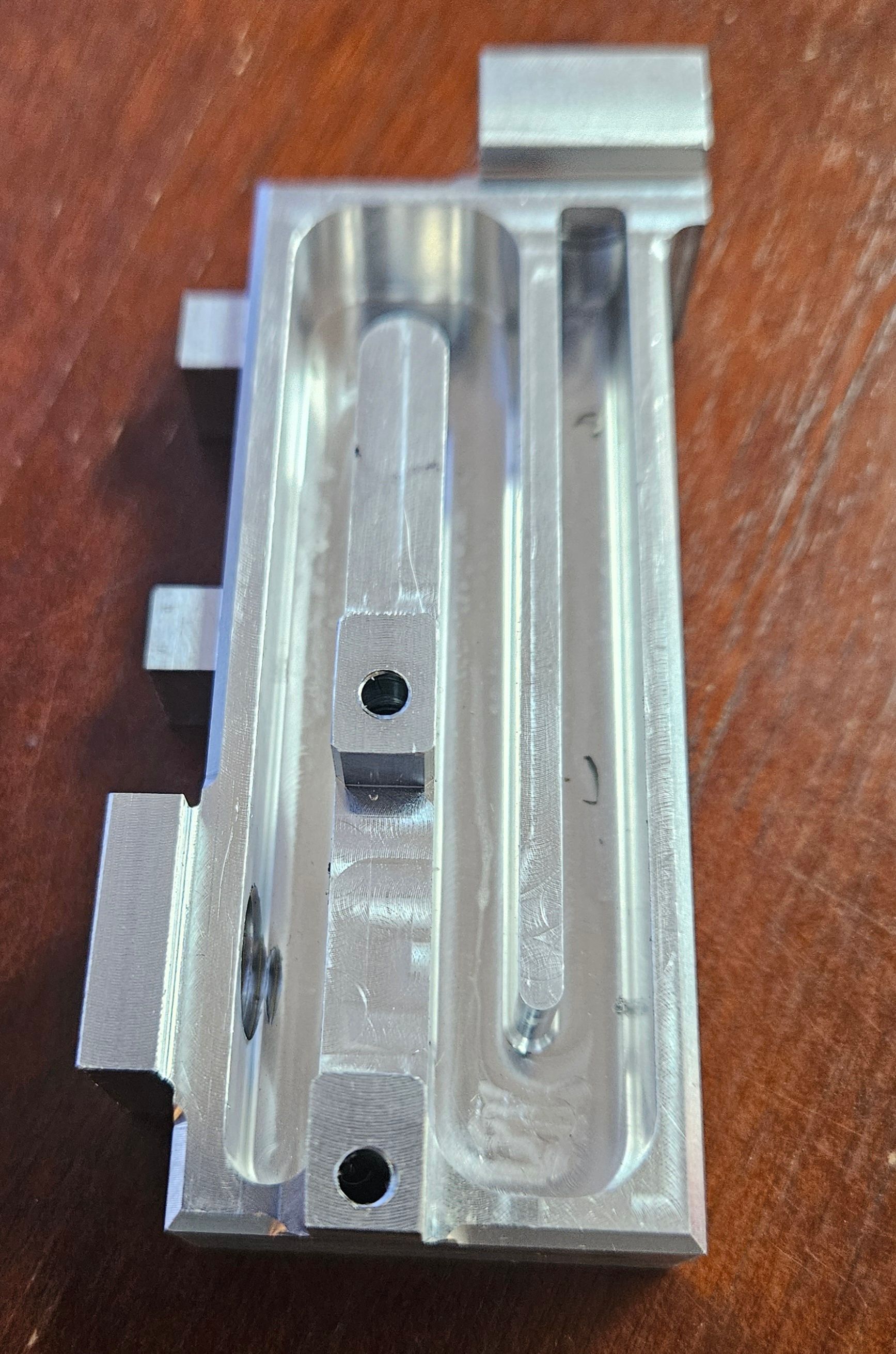 A rectangular metal component with CNC-machined grooves and holes, resting on a wooden surface.