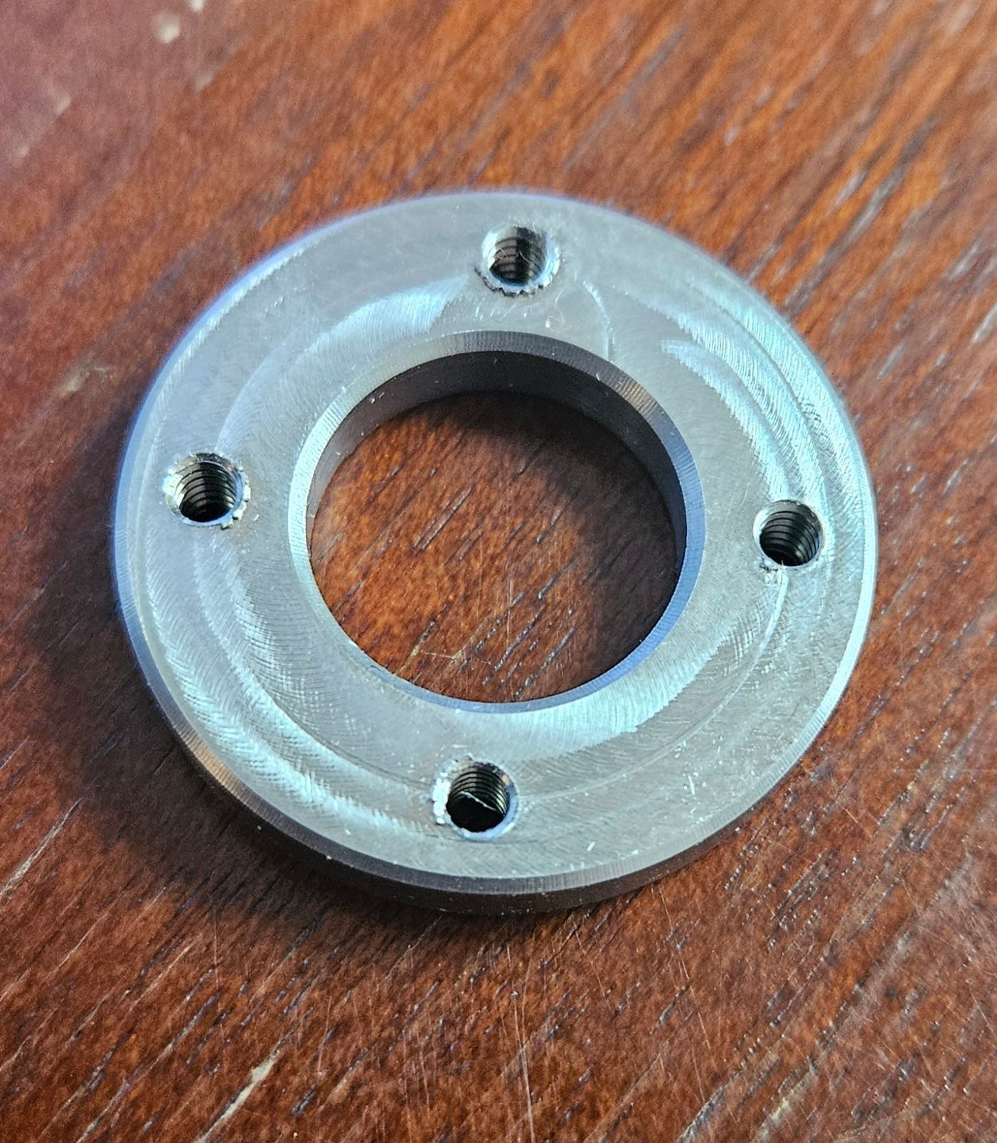 A circular metal washer with a central hole and four evenly spaced, threaded screw holes, resting on a wood surface.