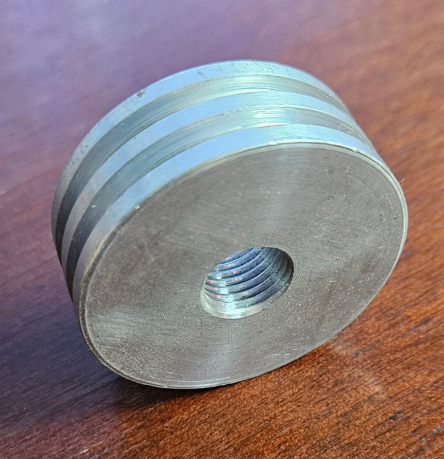 A small, circular, silver-colored metal spacer or washer with a threaded central hole resting on a wood-grain surface.
