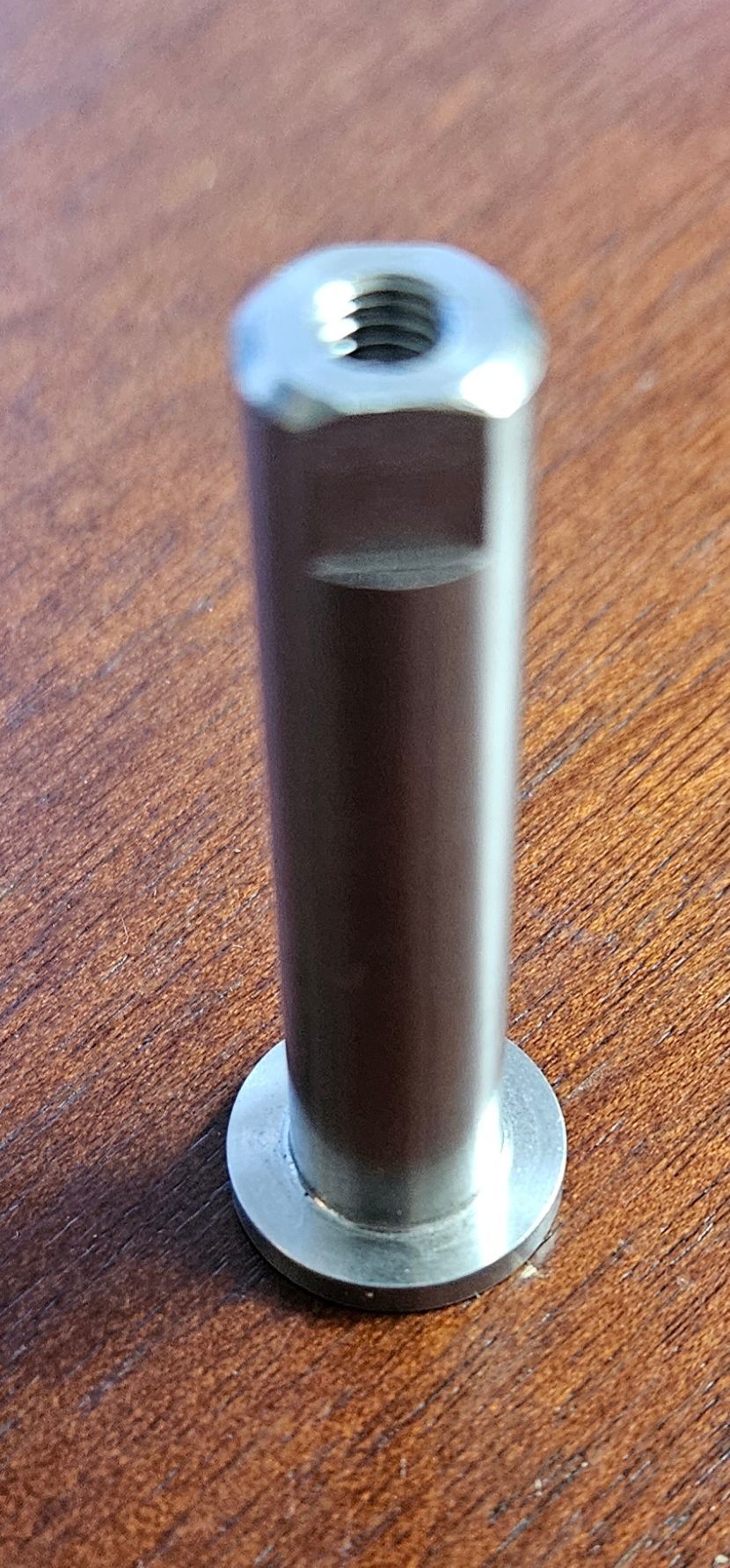 A silver-colored metal standoff spacer with a threaded hole at the top and a circular base, resting on a wood surface.