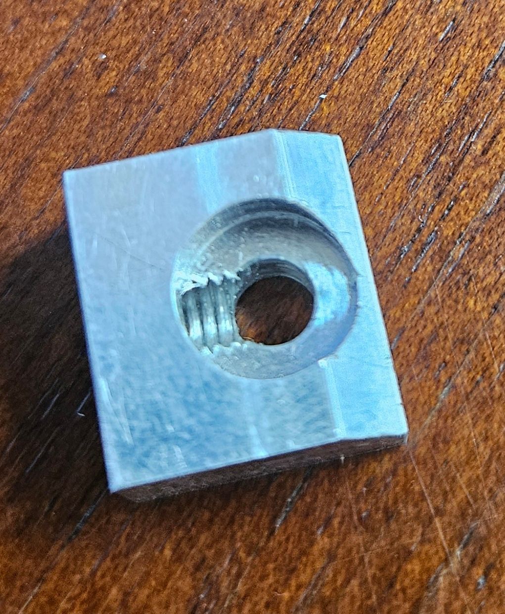A small, square, silver metal T-nut with a threaded hole, centered on a wooden surface.