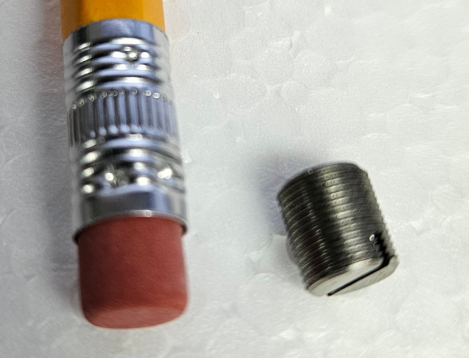 A pencil eraser next to a small, metal threaded insert with a slotted top, shown on a white styrofoam surface.