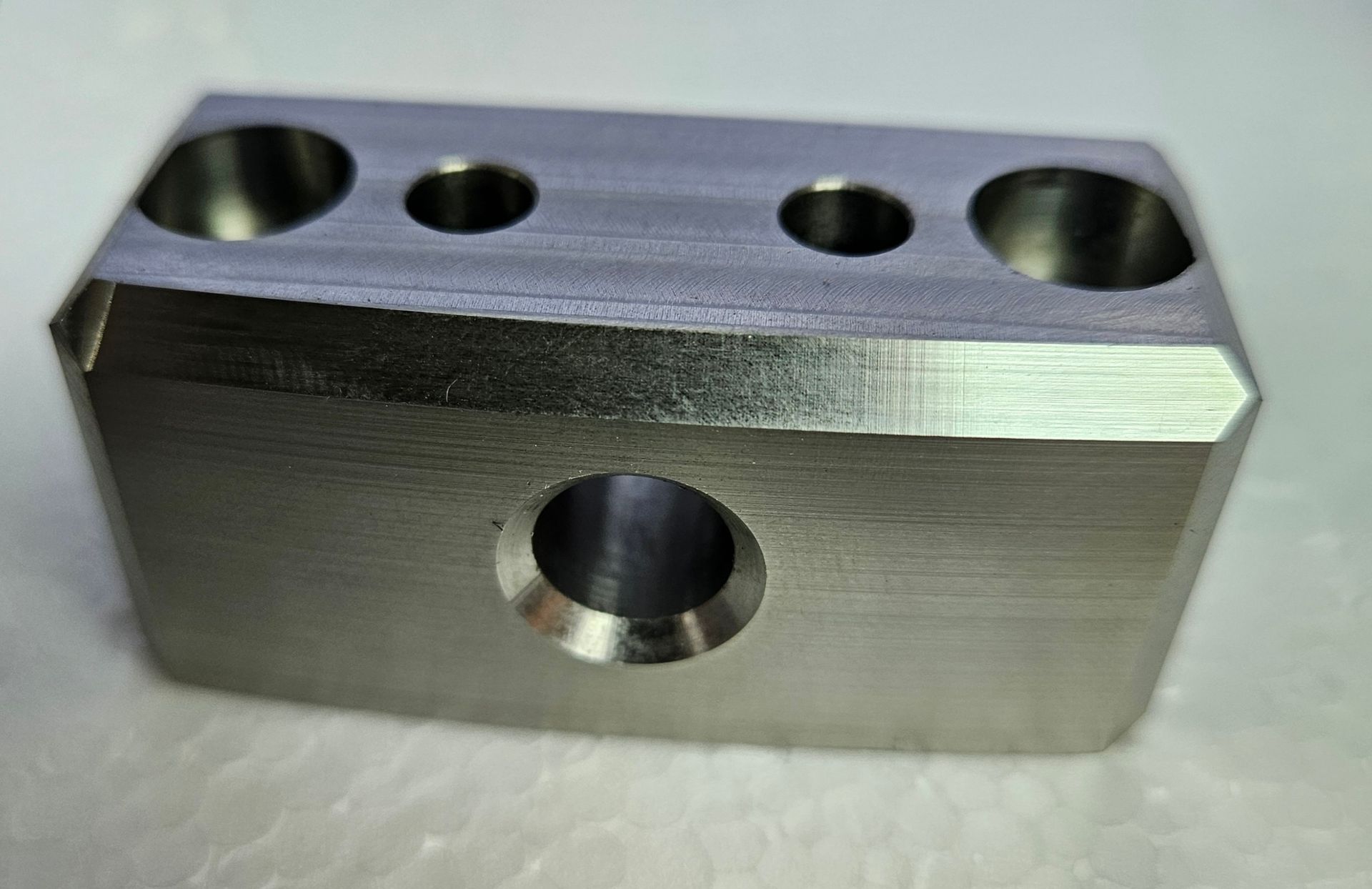A rectangular, metallic block with beveled edges, featuring one large central hole on the side and four holes on top.