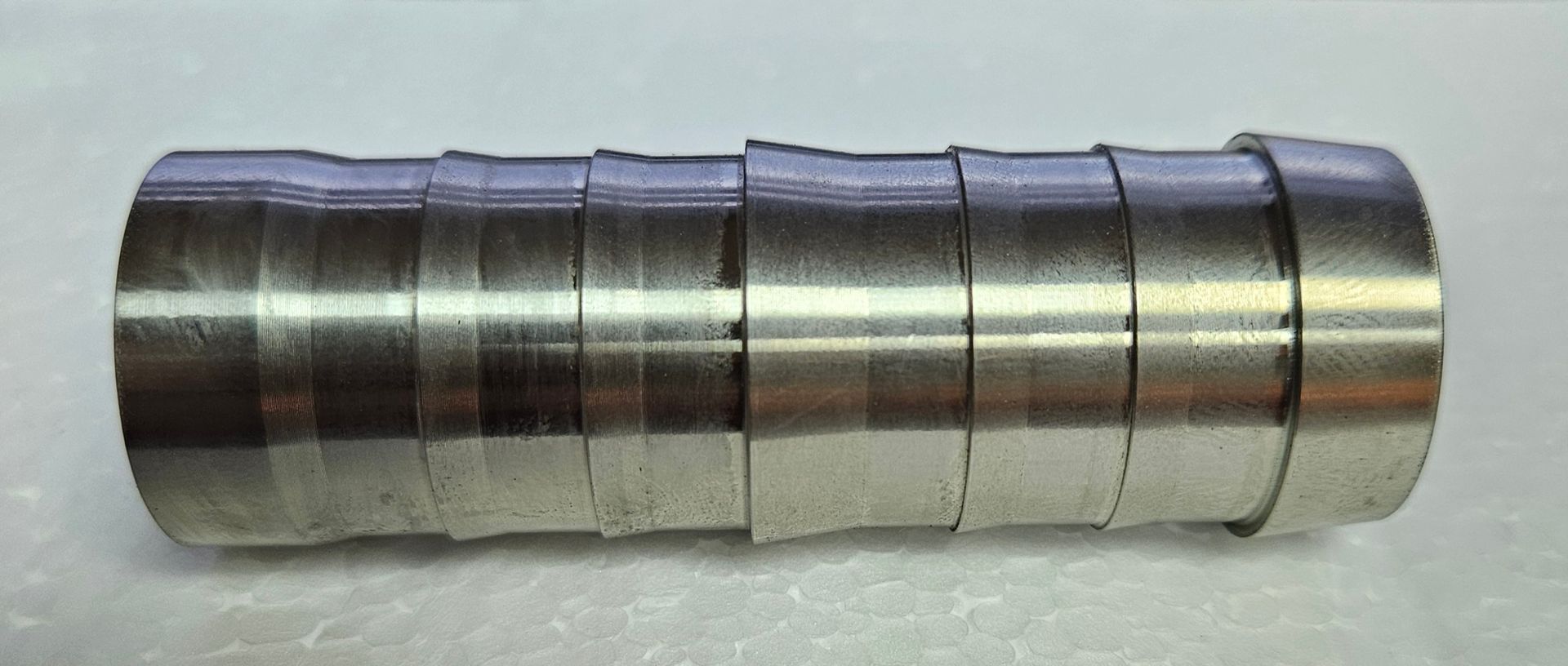 A silver-colored metal hose barb fitting with a ribbed design for secure tubing attachment, set against a white background.