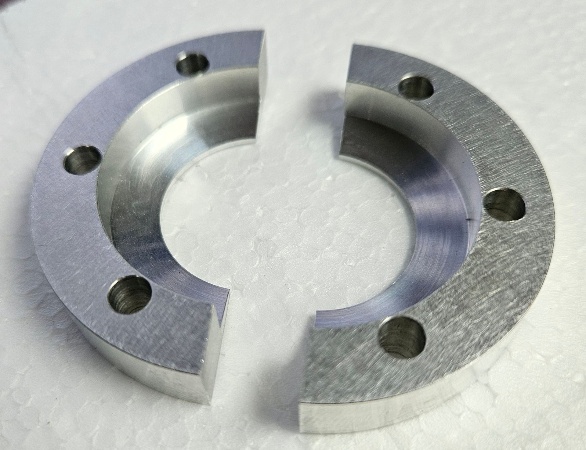 Two semi-circular, metallic, machined rings with bolt holes, lying on a white, textured surface.