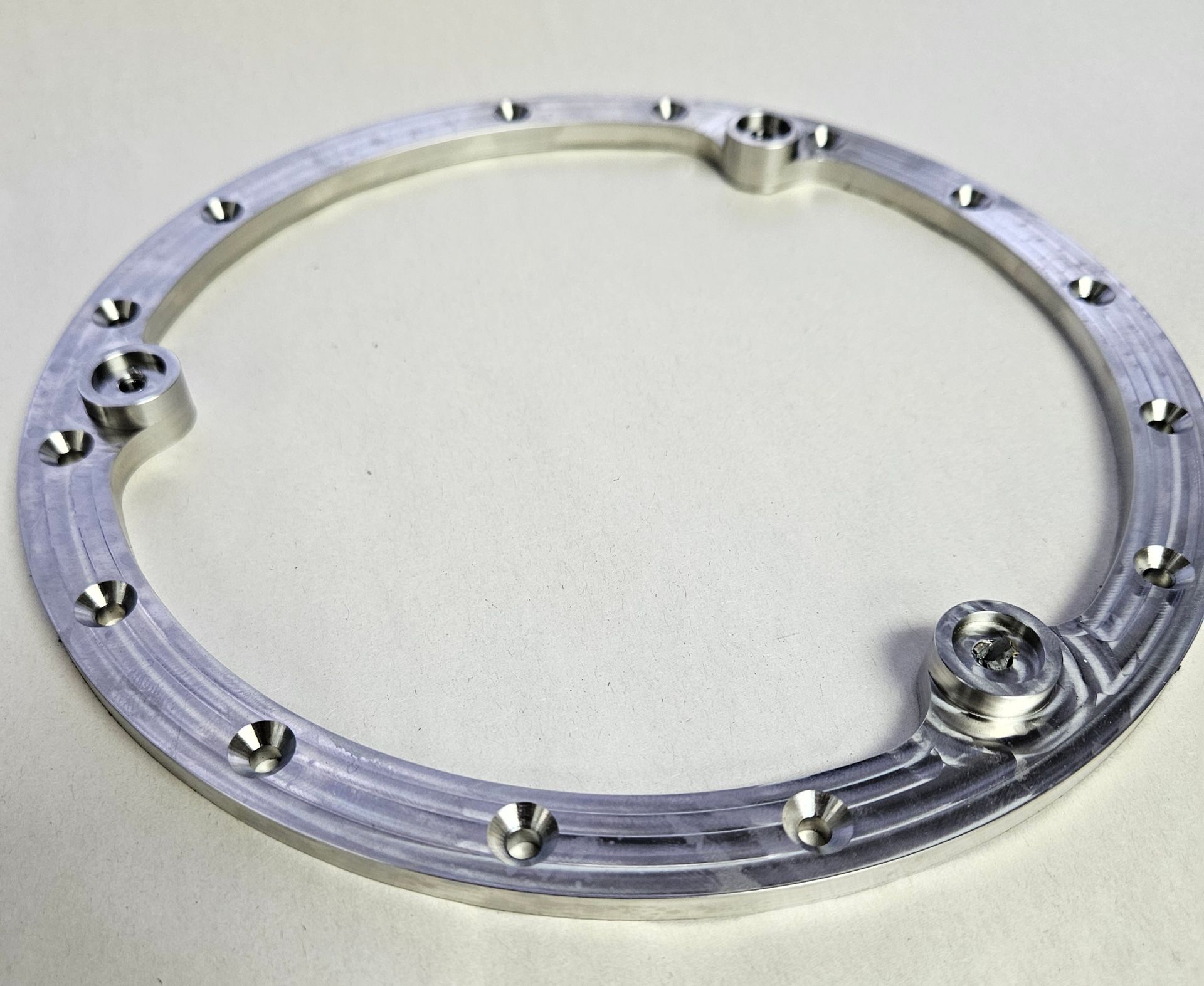 A silver-colored, circular metal ring with multiple countersunk screw holes and two raised cylindrical mounting points.