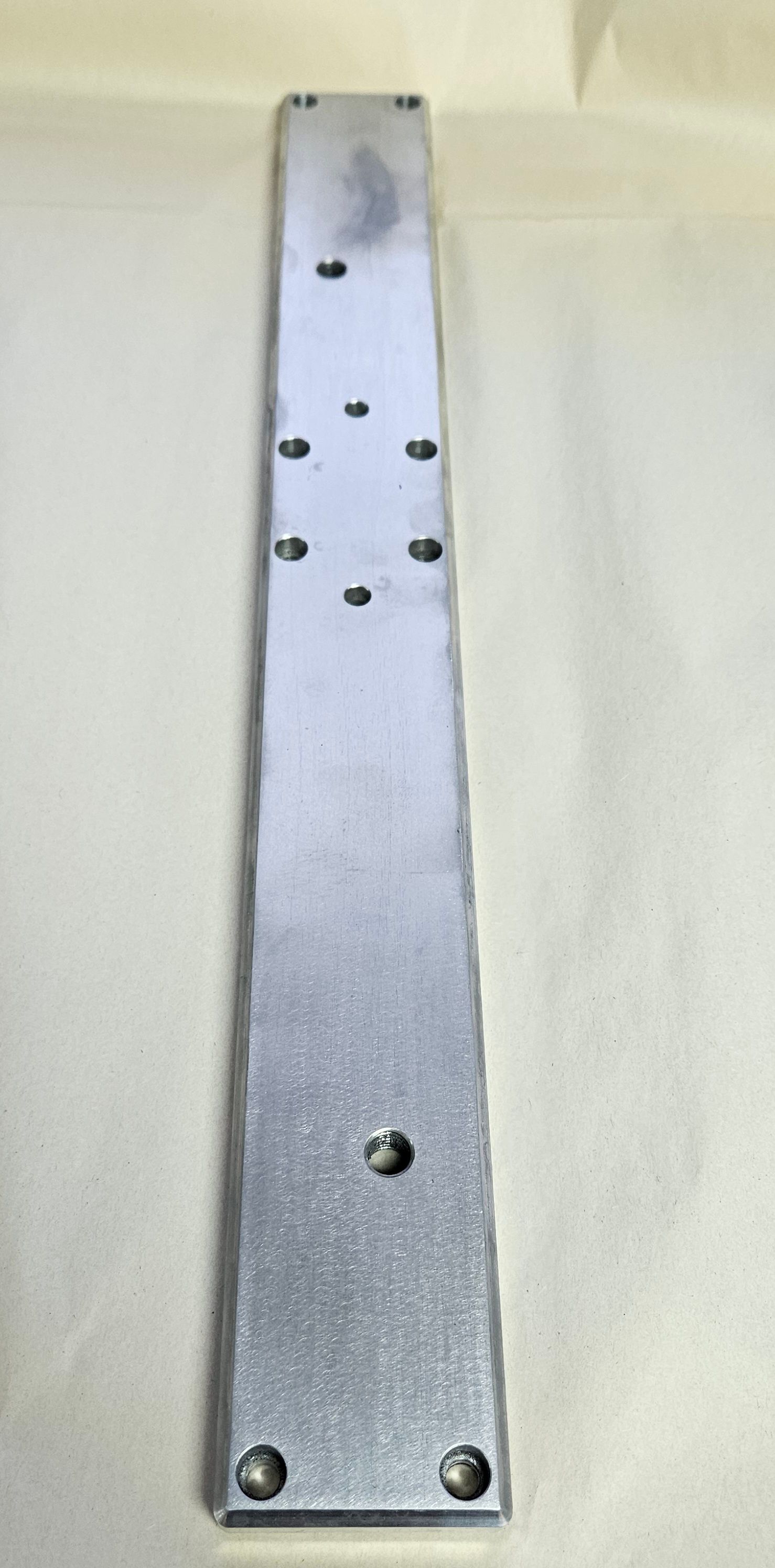 A long, rectangular, metallic bar featuring a symmetrical arrangement of drilled holes and a brushed silver finish.