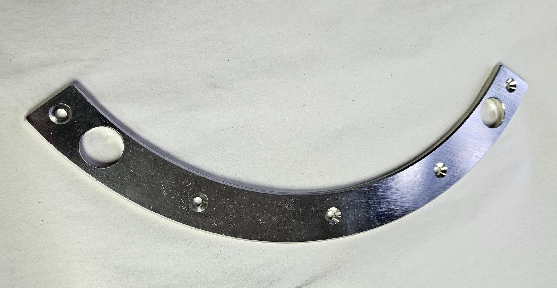 A metallic, arc-shaped metal bracket with a hole at each end and several small dimpled fasteners along its curve.
