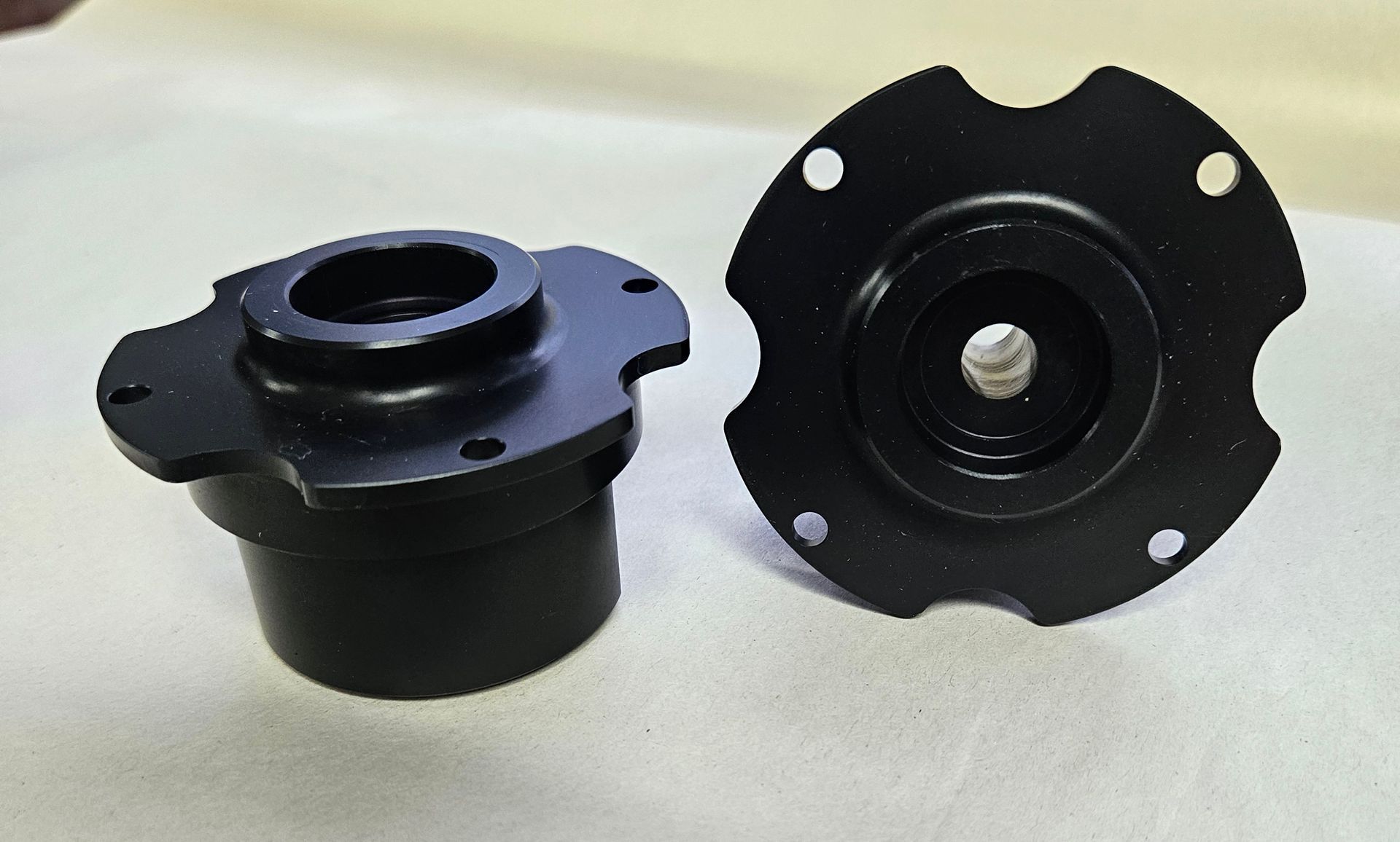 Two black metal flange components, one shown from the side and the other from above, resting on a white surface.