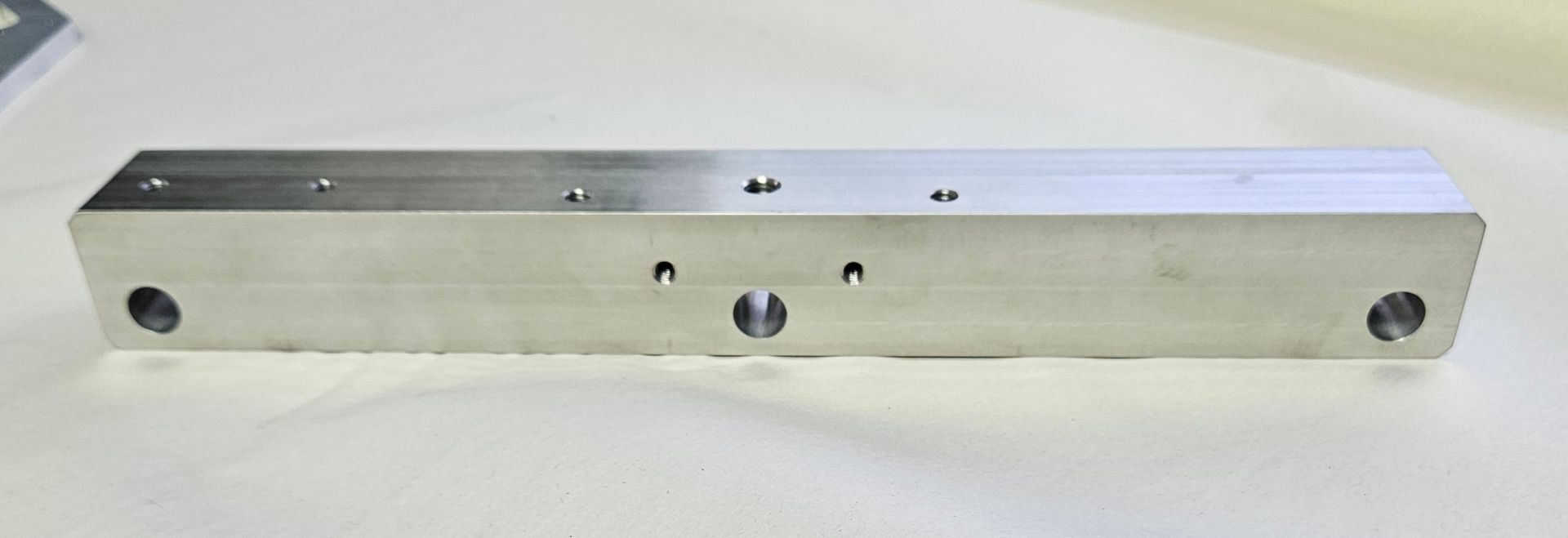 A long, rectangular, metallic industrial bar with various drilled holes and ports, set against a plain light background.